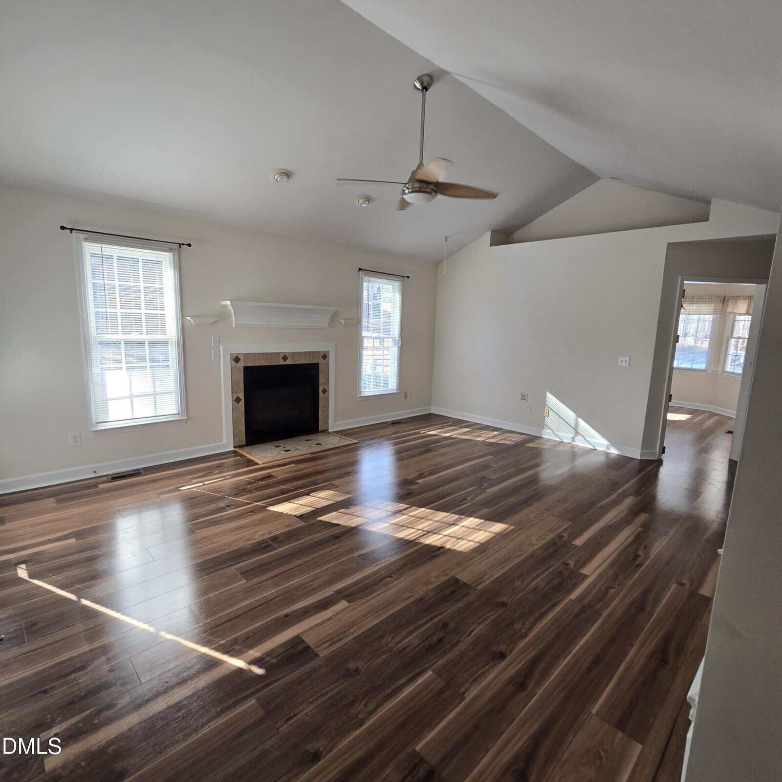 Spacious, empty living room with wood floors, a ceiling fan, two large windows, and a fireplace. Sunlight streams in, casting shadows on the floor. Walls are painted light beige.
