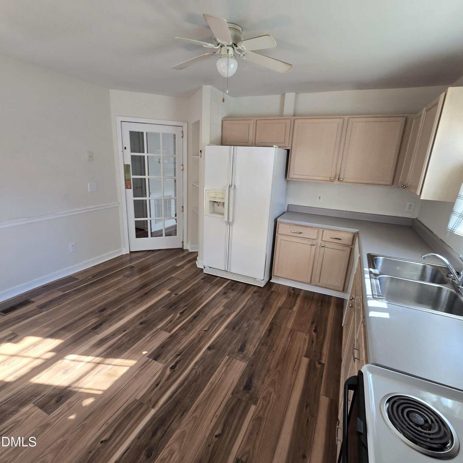 A kitchen with wood flooring, light-colored cabinets, a white refrigerator, a white stove, a stainless steel sink, and a ceiling fan. A glass-paneled door is on the left side of the room.