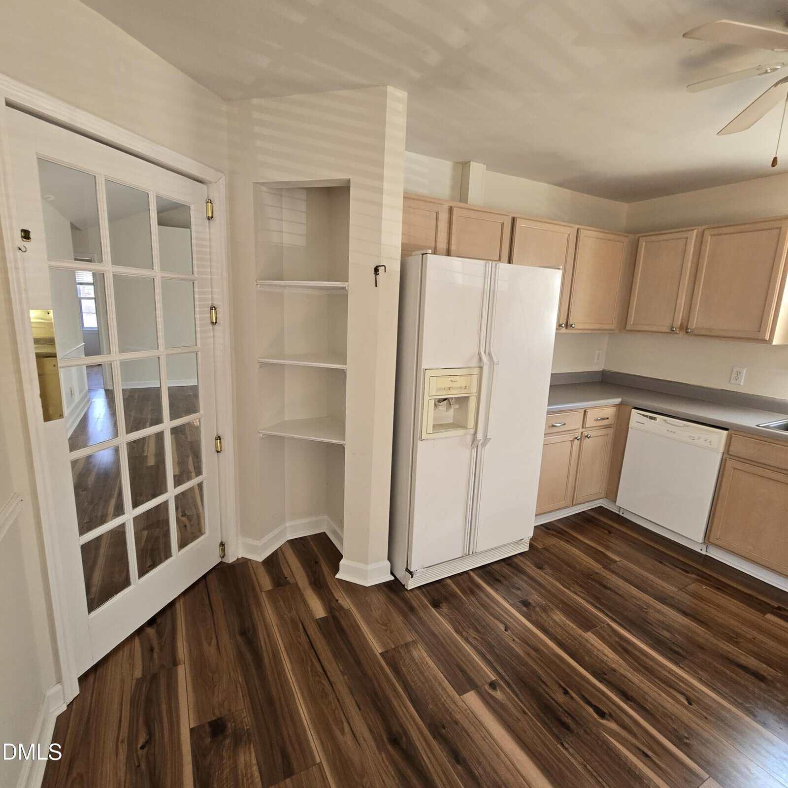 A kitchen with wood floors, light wood cabinets, white countertops, a white refrigerator, a white dishwasher, built-in corner shelves, and a glass-paned door. The room appears clean and well-lit.