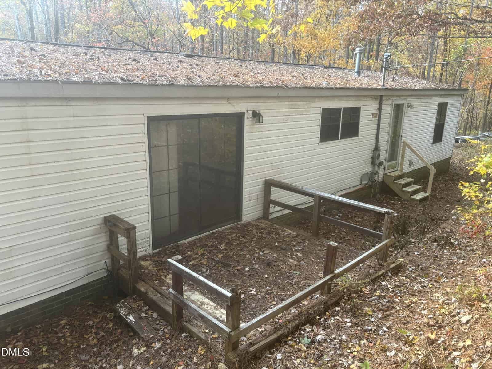 A white manufactured home with a sliding glass door, small wooden ramp, and steps leading to a wooded yard covered in fallen leaves during autumn.
