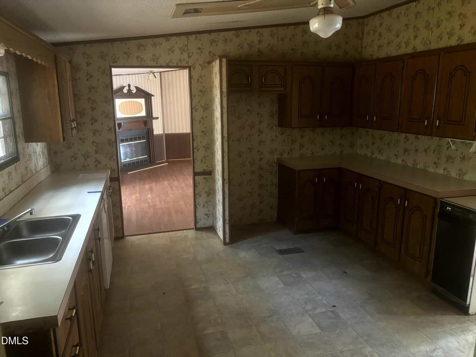 A dated kitchen with brown wood cabinets, beige countertops, floral wallpaper, a double sink under a window, dishwasher, ceiling fan, and an open doorway leading to a living area with wood flooring.
