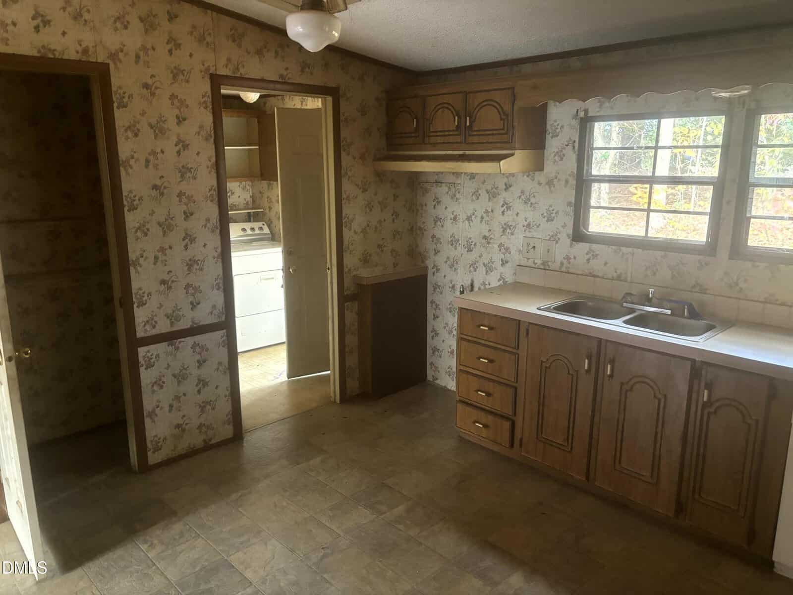 A vintage kitchen with floral wallpaper, wood cabinets, a double sink under two windows, and beige tile floors. An open door reveals a laundry room with a washer and dryer.
