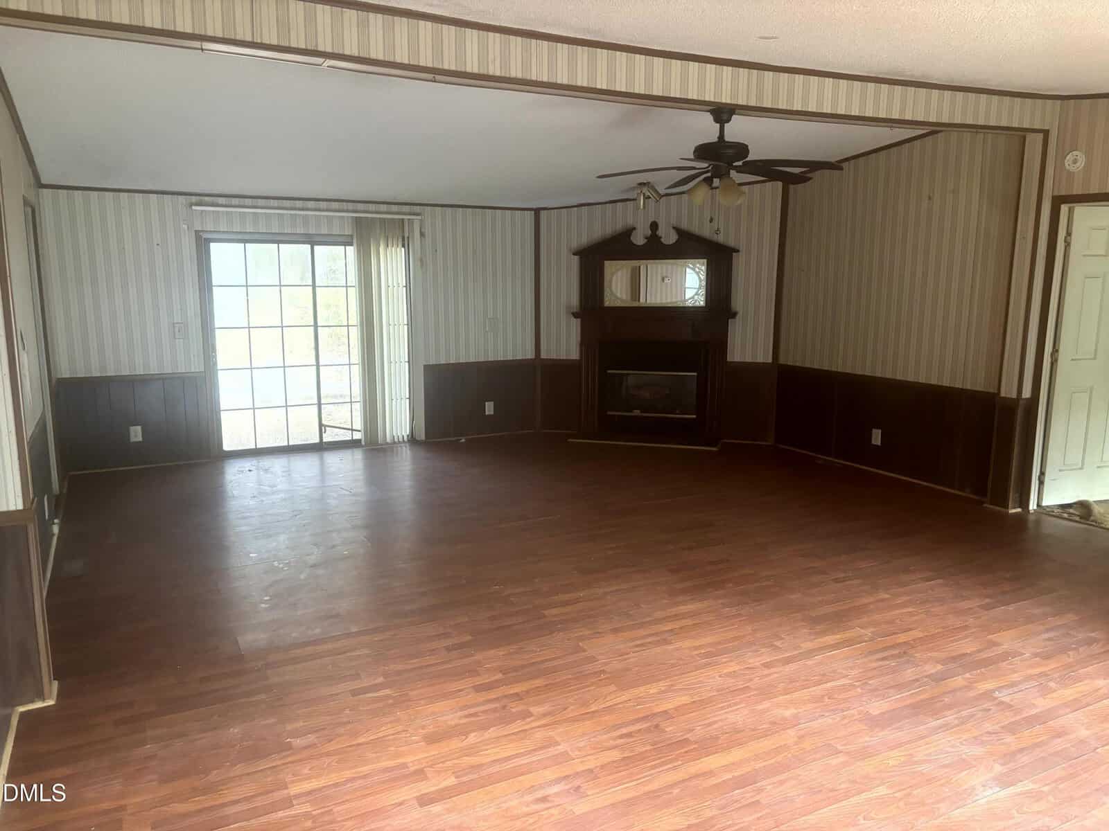 Empty living room with brown wood floors, striped wallpaper, a ceiling fan with light, a fireplace with a mirror above it, sliding glass doors on one wall, and partial wood paneling on the walls.