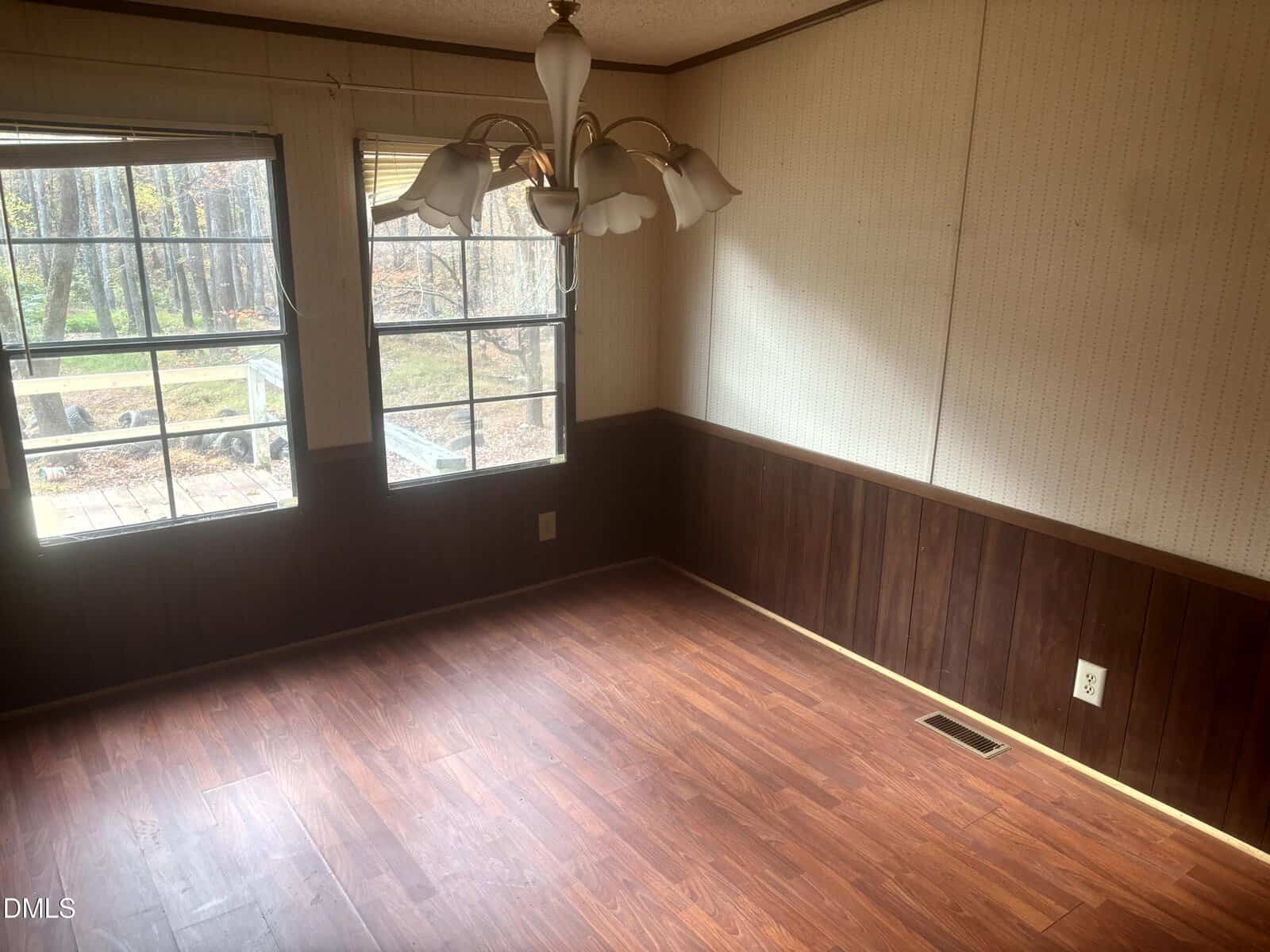 Empty room with wood-style laminate flooring, dark wood paneling on the lower walls, light wallpaper on the upper walls, a small chandelier-style light fixture, and two windows showing an outdoor view.
