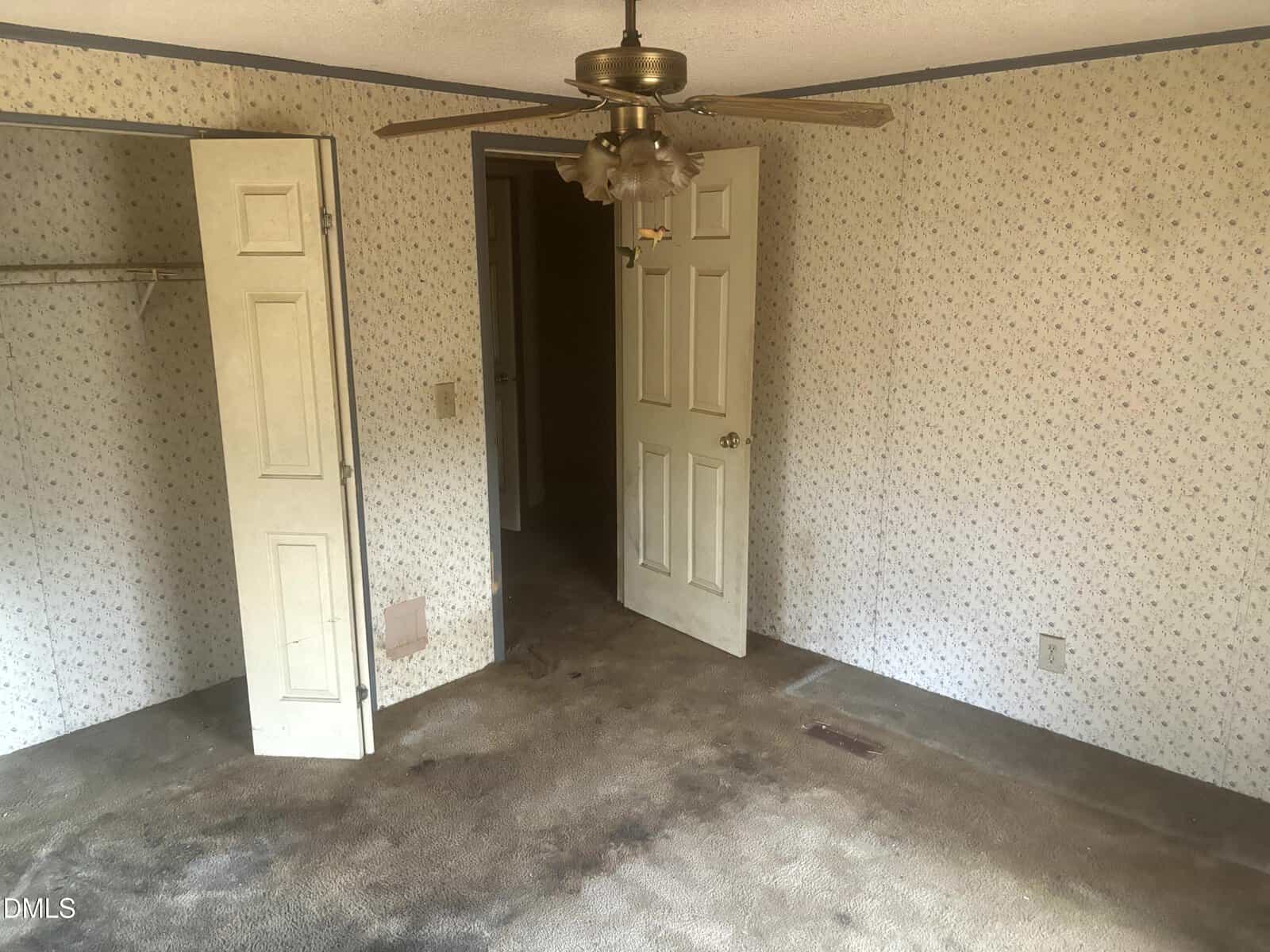 A small, empty room with old floral wallpaper, stained brown carpet, an open closet, an open door, and a ceiling fan with a light. There is visible wear on the closet and carpet.