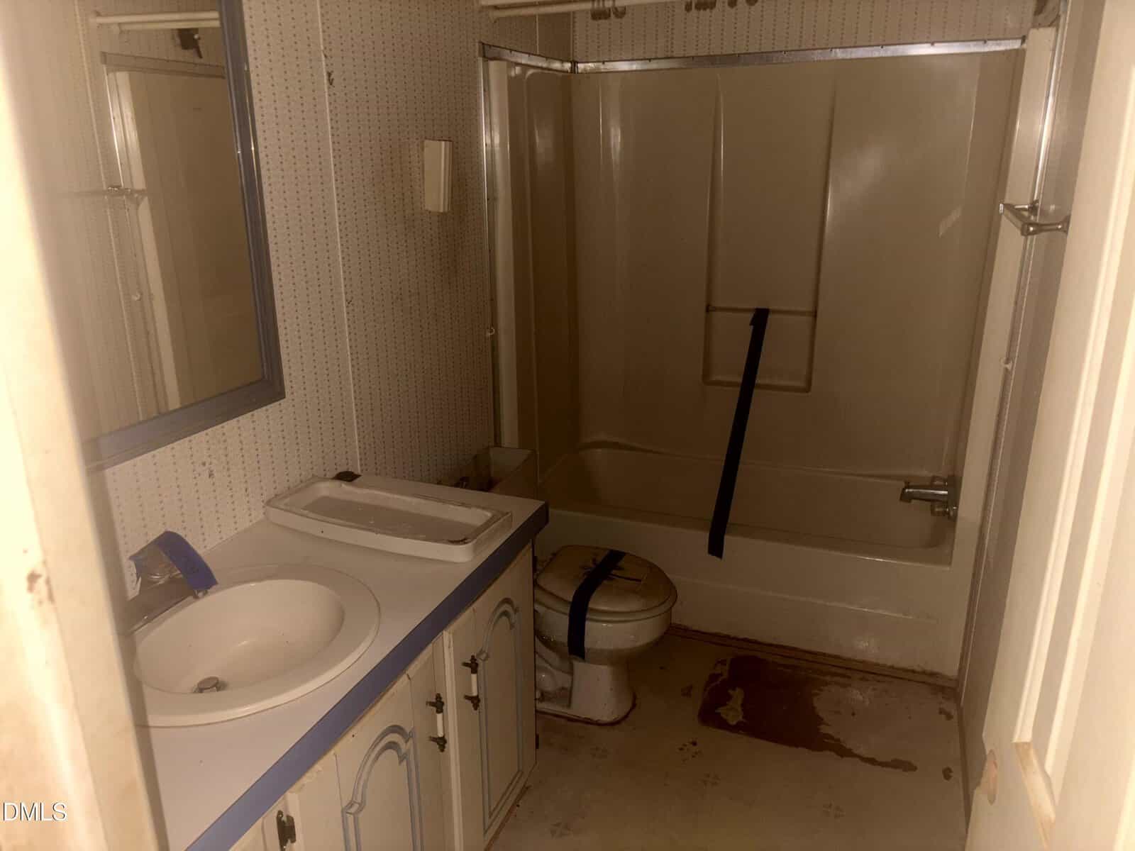 A bathroom with a bathtub-shower combo, toilet, and sink. The toilet and tub are taped shut, and there are stains on the floor. The room has wallpaper, a mirror, and worn cabinets. The lighting is dim.