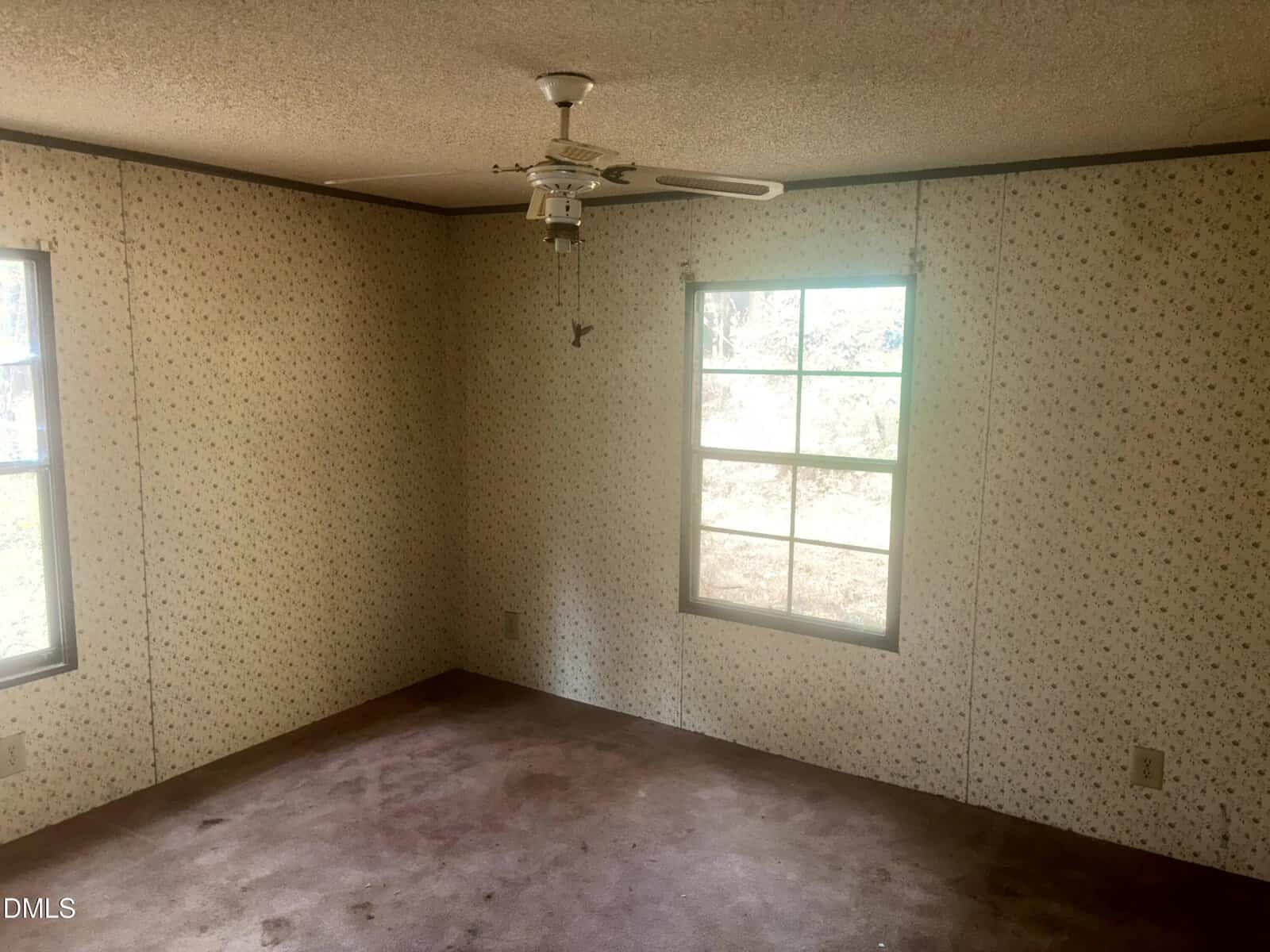 Empty room with two windows, floral-patterned wallpaper, a ceiling fan with light fixture, and worn carpet. Sunlight is coming through the windows, illuminating part of the floor and walls.