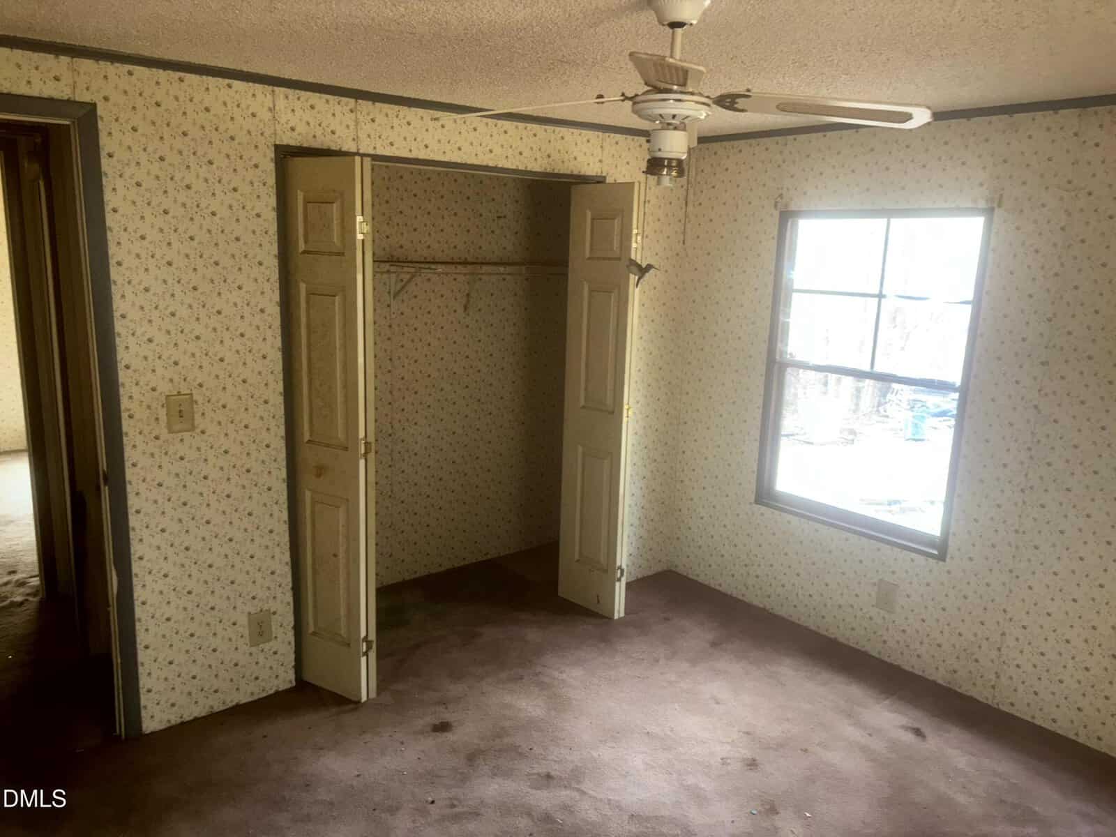 A small empty room with floral wallpaper, a ceiling fan, an open closet with double doors, a window letting in daylight, and worn carpet flooring. An open doorway leads to another room.