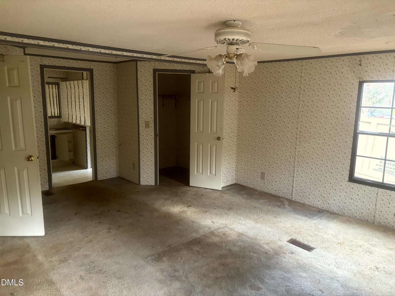 A sparsely furnished, carpeted room with stained flooring, wallpapered walls, a ceiling fan with broken blades, two open doors leading to other rooms, a window, and visible natural light.