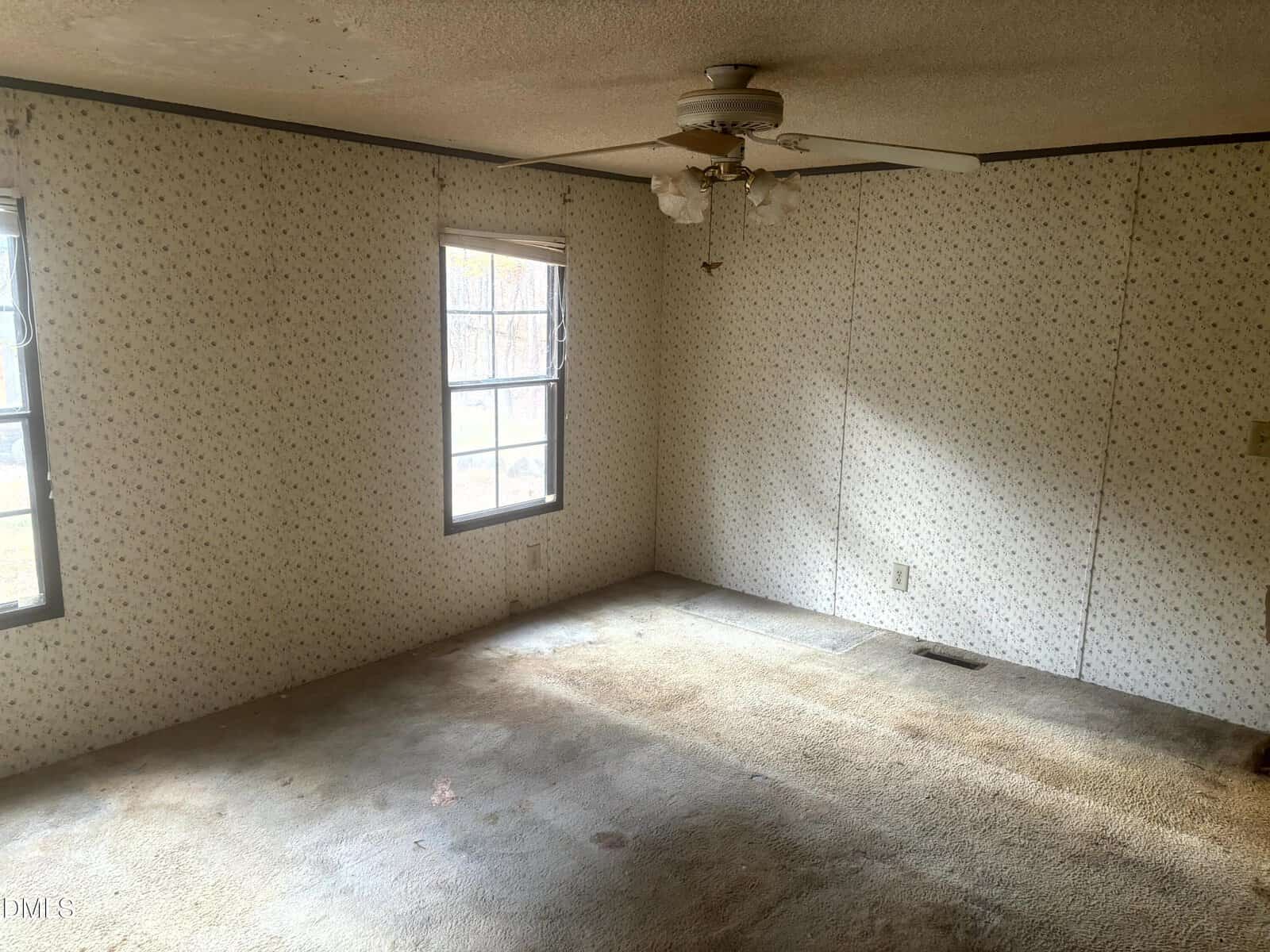 A small, empty room with two windows, stained carpet, patterned wallpaper, and a ceiling fan with a missing or broken blade. Sunlight comes in through the windows, highlighting the worn condition of the space.