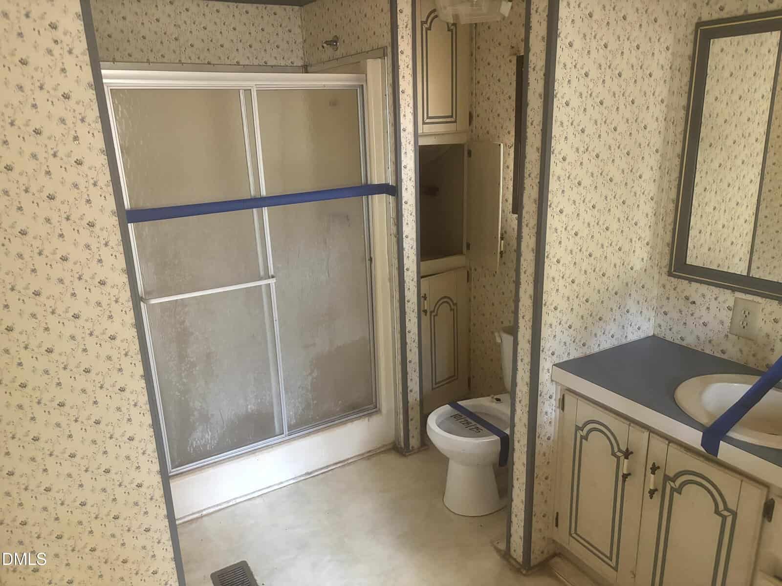 A bathroom with floral wallpaper, a shower with sliding doors, a toilet, cabinets, and a sink. Blue tape is across the shower doors and toilet, indicating they are not in use.