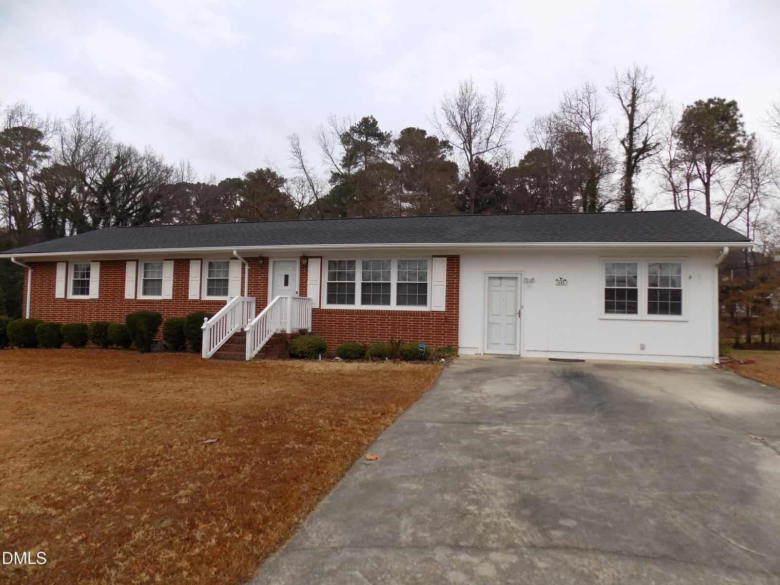 Single-story house with red brick and white siding, black shingle roof, white front steps, shrubbery along the front, and a concrete driveway leading to a garage. Leafless trees and brown grass surround the home.