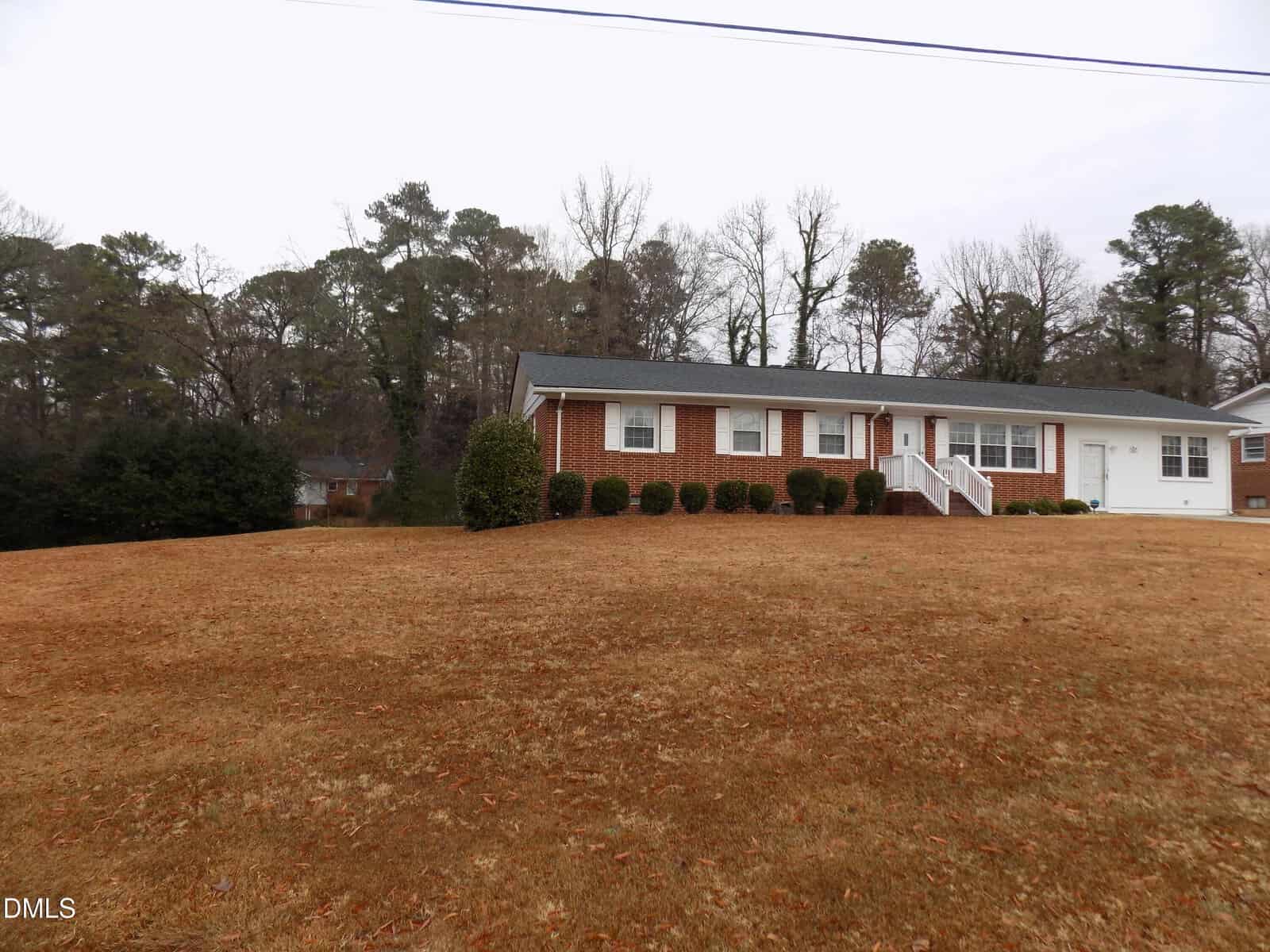 Single-story brick house with white trim, front porch, and a row of shrubs. The house sits on a large, sloped, brown grass lawn with trees and neighboring houses visible in the background.