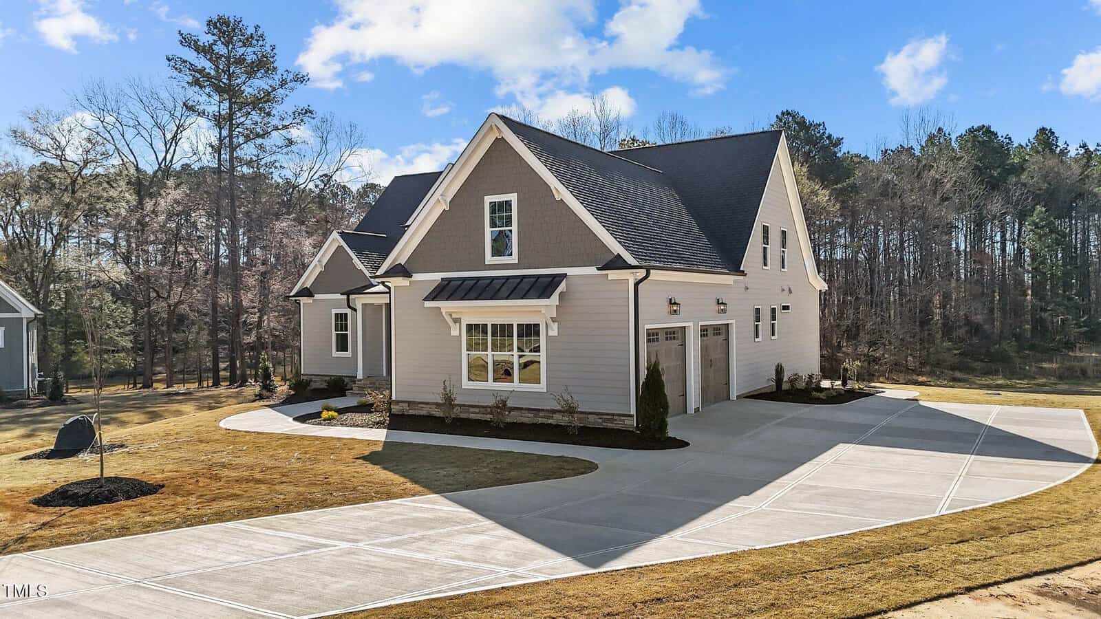 A modern two-story house with gray siding, a dark sloped roof, and white trim, featuring a double garage and a large concrete driveway surrounded by a lawn and trees in the background.