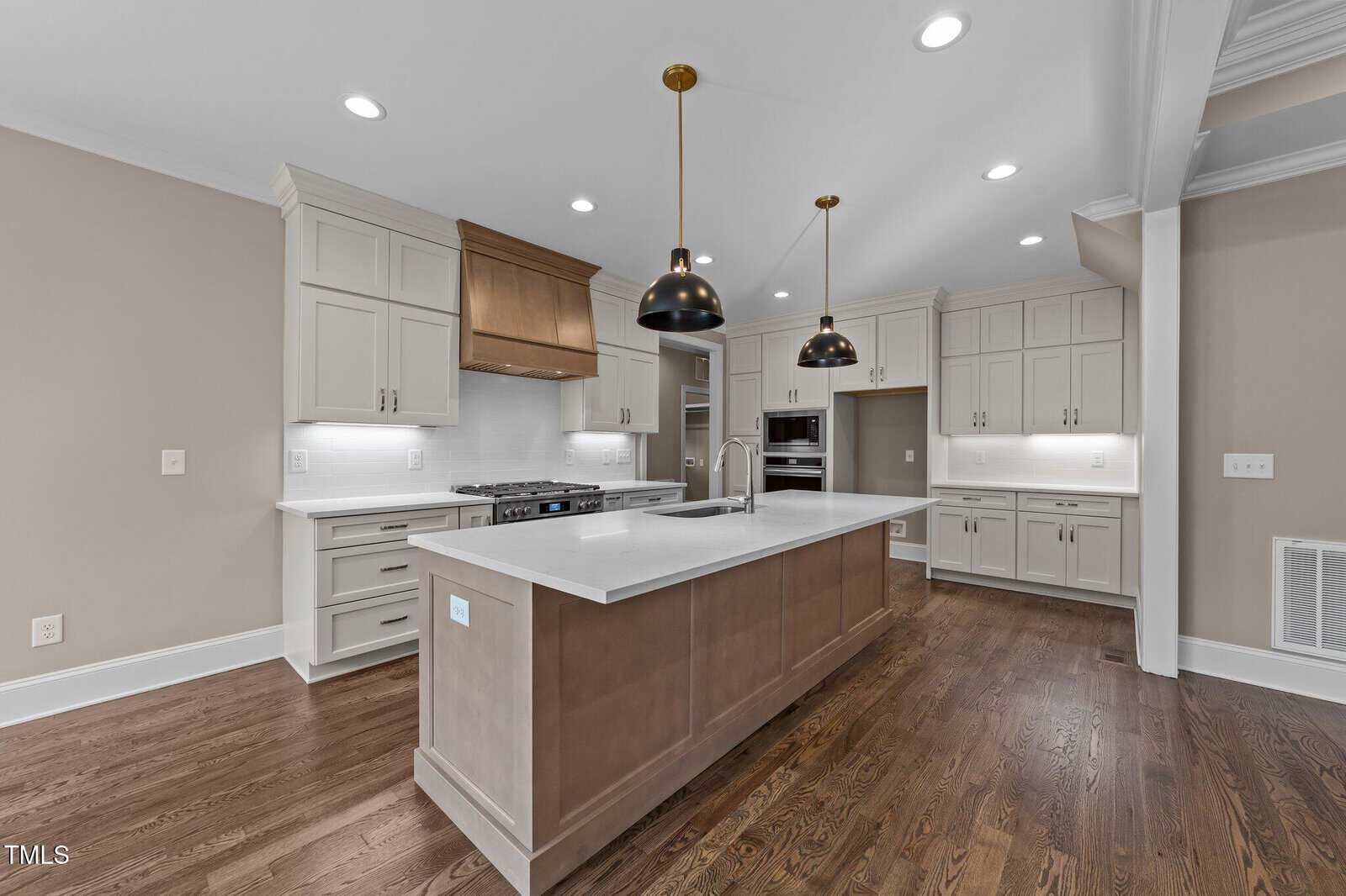 Modern kitchen with a large central island, white cabinetry, stainless steel appliances, wooden floors, two black pendant lights above the island, and neutral-colored walls.