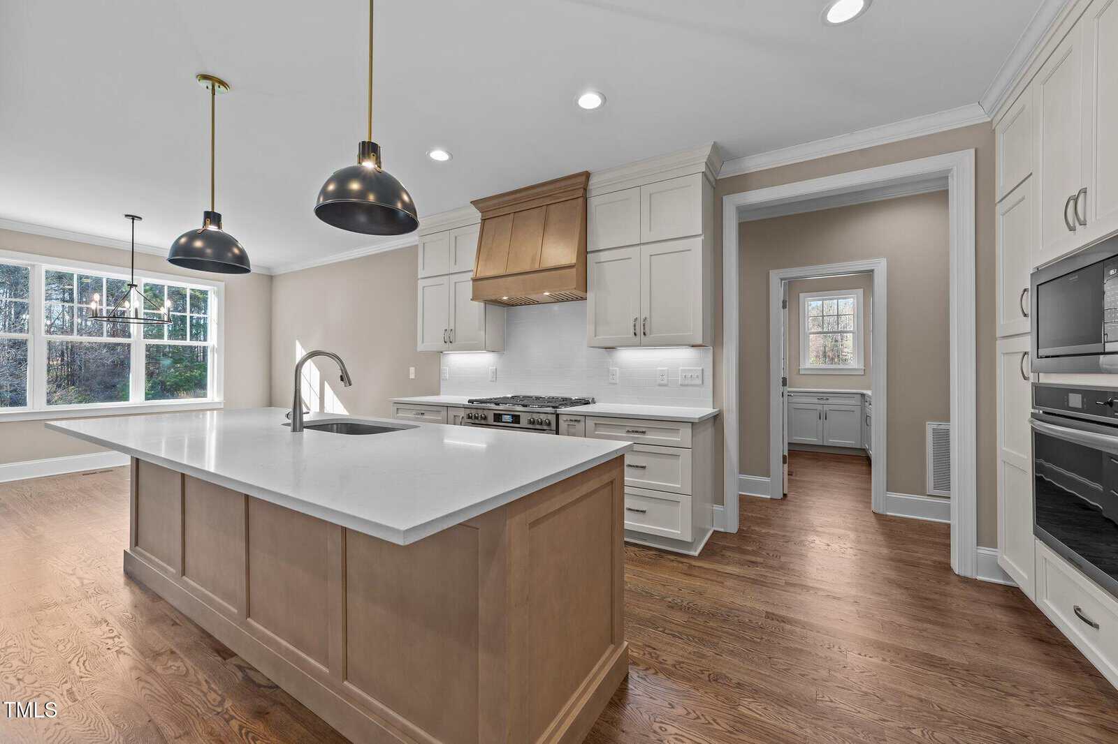 Modern kitchen with a large island, white quartz countertops, light wood floors, white cabinetry, stainless steel appliances, and black pendant lights. A window provides natural light, and a doorway leads to an adjacent room.
