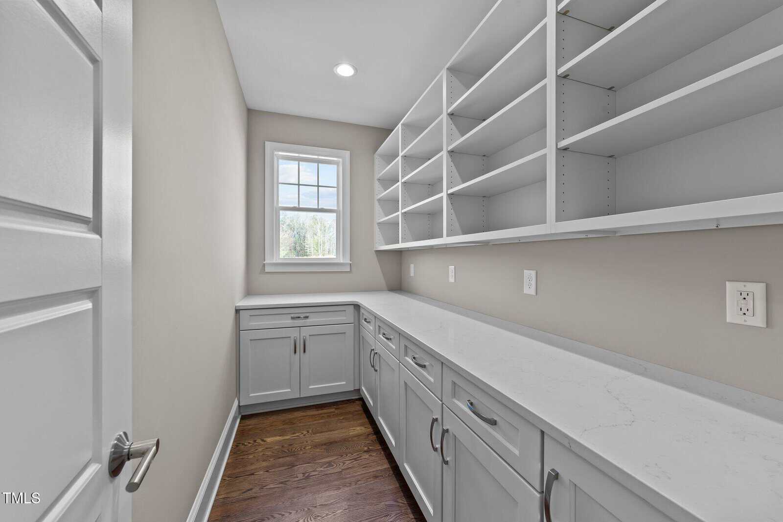 A narrow pantry room with white shelves, white cabinets, and a marble countertop. There is a window at the far end, and the floor is dark wood. The walls are light beige and the room is well-lit.