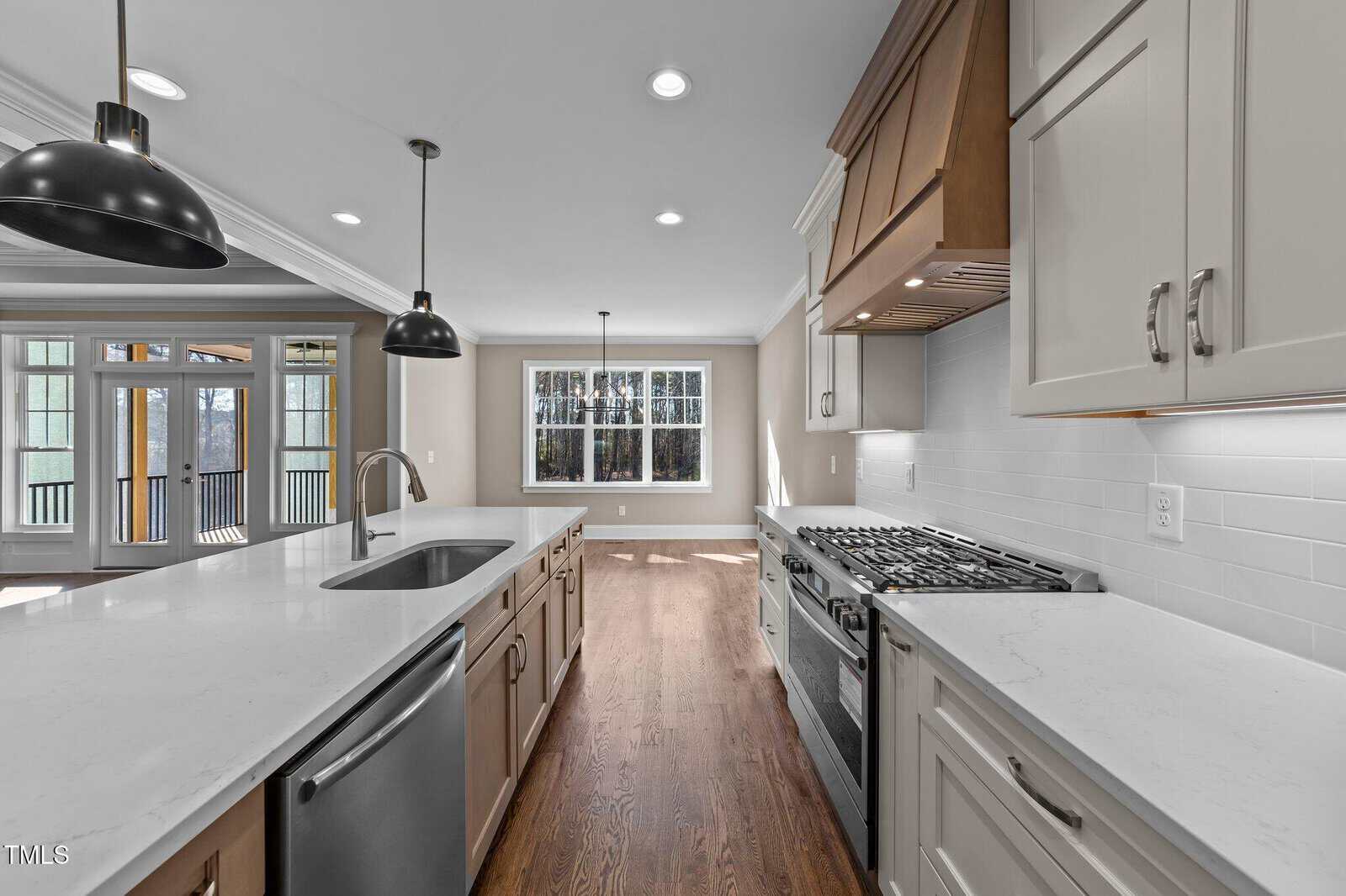 A modern kitchen with white cabinets, stainless steel appliances, a gas stove, large island with a sink, pendant lighting, hardwood floors, and a view of a dining area and deck through wide windows and glass doors.