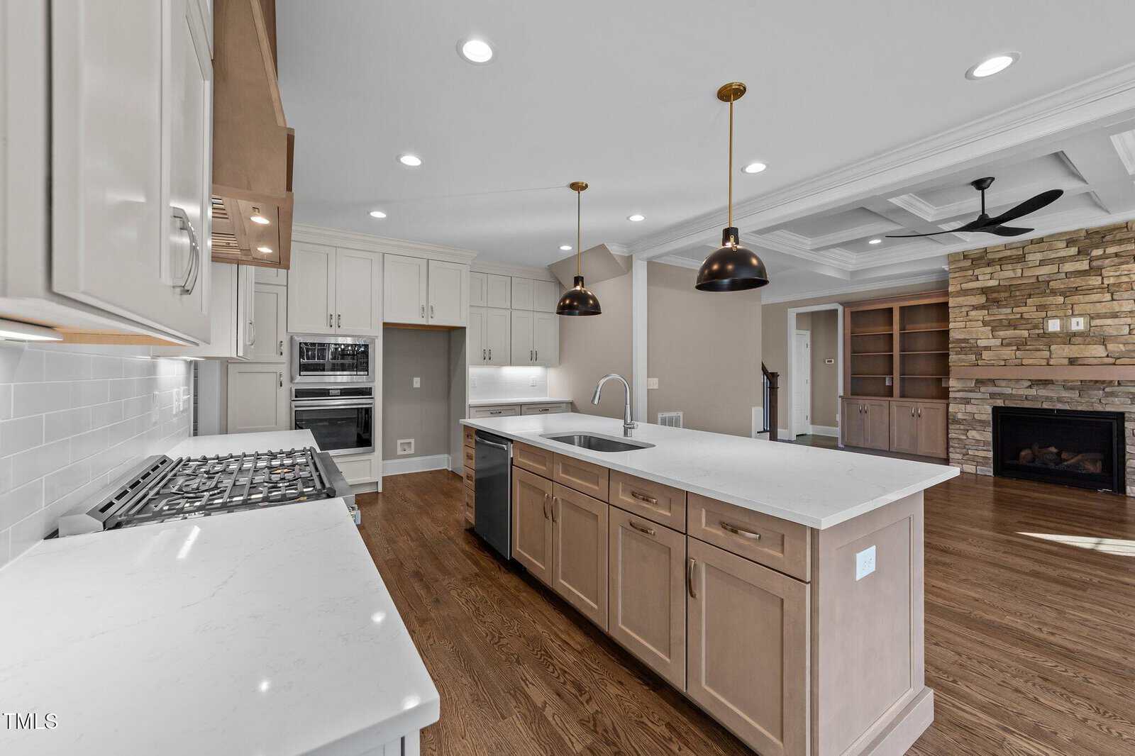 Modern kitchen with white cabinets, light wood island, white countertops, stainless steel appliances, pendant lights, hardwood floors, and an open living area with built-in shelves, fireplace, and ceiling fan in the background.