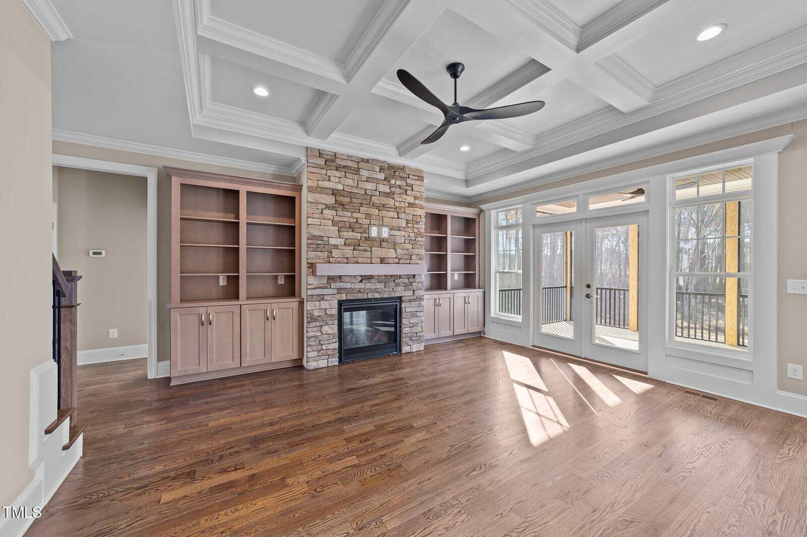 A bright living room with hardwood floors, built-in shelves flanking a stone fireplace, a ceiling fan, coffered ceiling, and large windows with glass doors opening to a deck with wooded views.