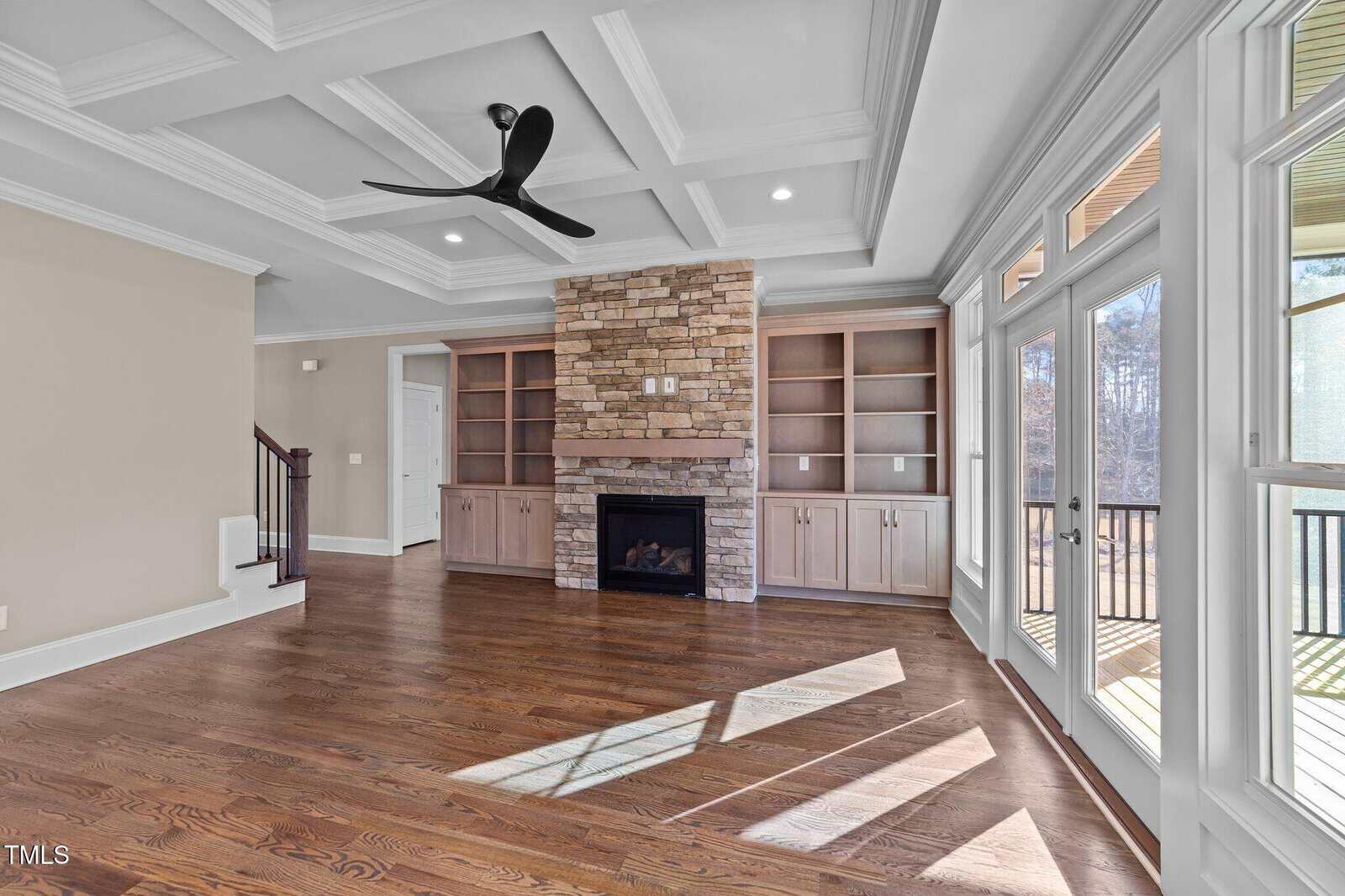 A spacious, sunlit living room with hardwood floors, a coffered ceiling, a modern black ceiling fan, stone fireplace flanked by built-in shelves, and large windows with a door leading to a deck outside.