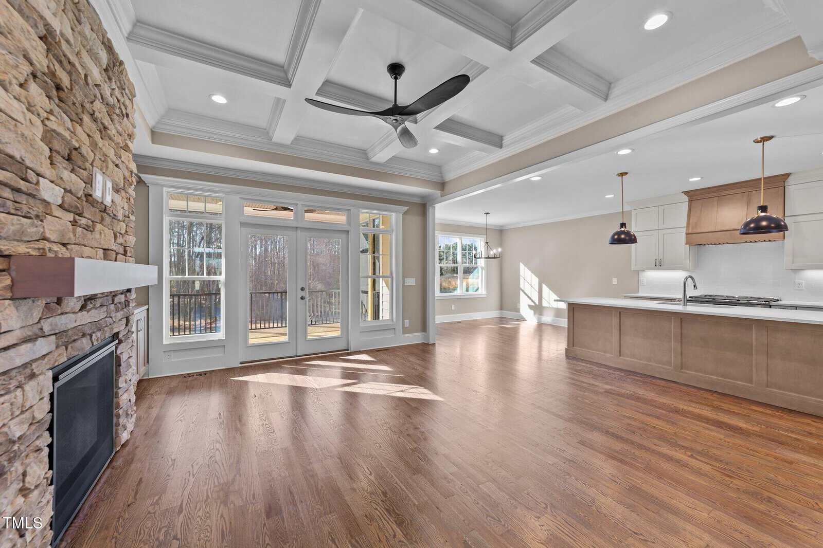 Spacious open-concept living area with hardwood floors, a stone fireplace, coffered ceiling, ceiling fan, large windows, and an adjacent modern kitchen with pendant lights and an island.