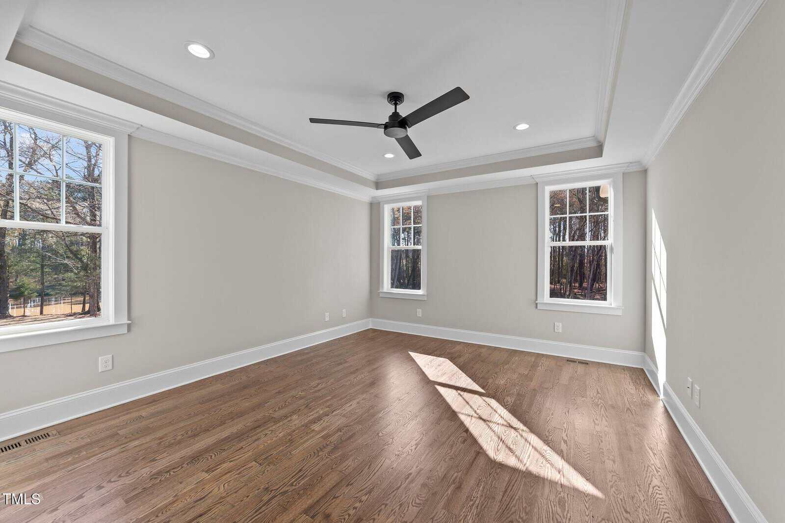 Empty room with light beige walls, wood flooring, tray ceiling with recessed lighting, black ceiling fan, and two large windows letting in natural light; there are trees visible outside the windows.