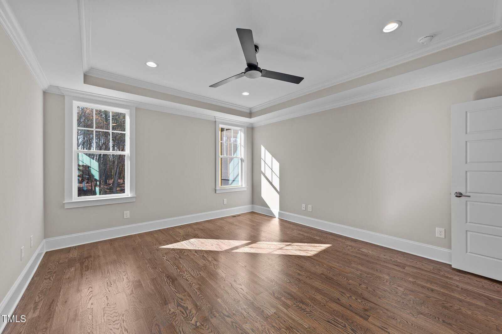 Empty room with hardwood floors, beige walls, white trim, two windows, a ceiling fan, recessed lighting, and a white paneled door. Sunlight shines through one window, creating a bright spot on the floor.