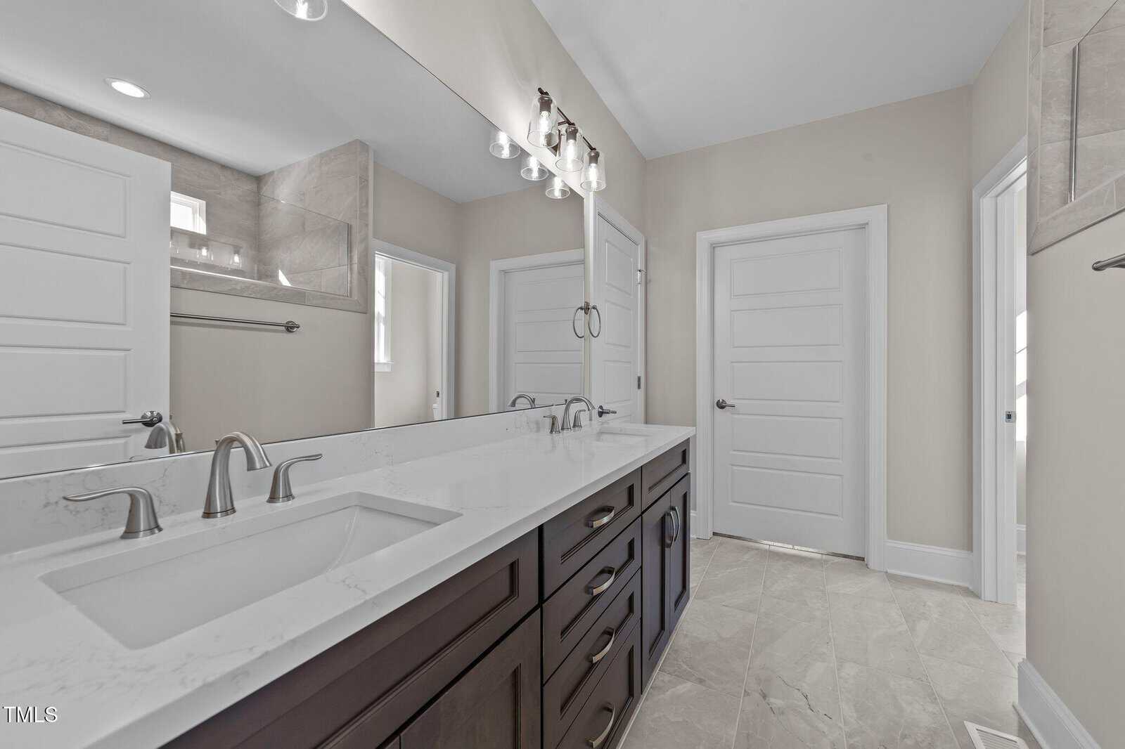 Modern bathroom with light beige walls, a double vanity featuring a white countertop, two sinks with chrome fixtures, a large mirror, and tile flooring. There are two white paneled doors and a glass shower enclosure.