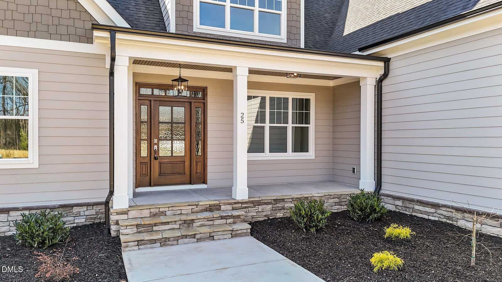 Front entryway of a house with a wooden door, sidelights, and a hanging porch light. The porch has white columns, a stone base, and is surrounded by mulch and small shrubs. House siding is light gray.