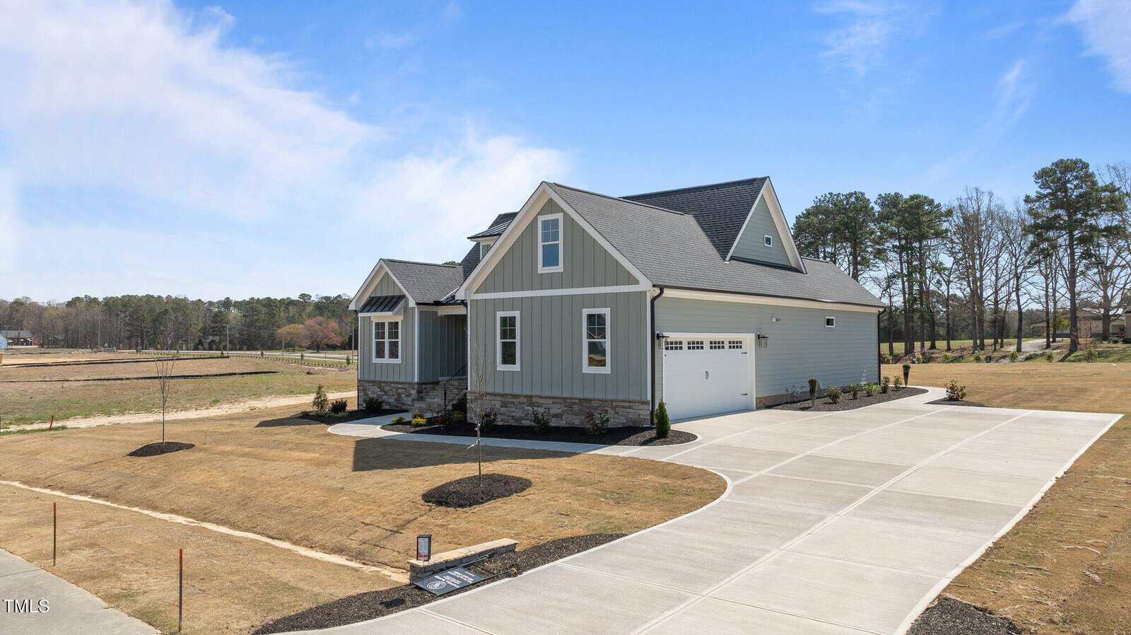 A modern two-story house with gray siding, stone accents, and a two-car garage sits on a large lot with a wide concrete driveway and minimal landscaping under a blue sky.