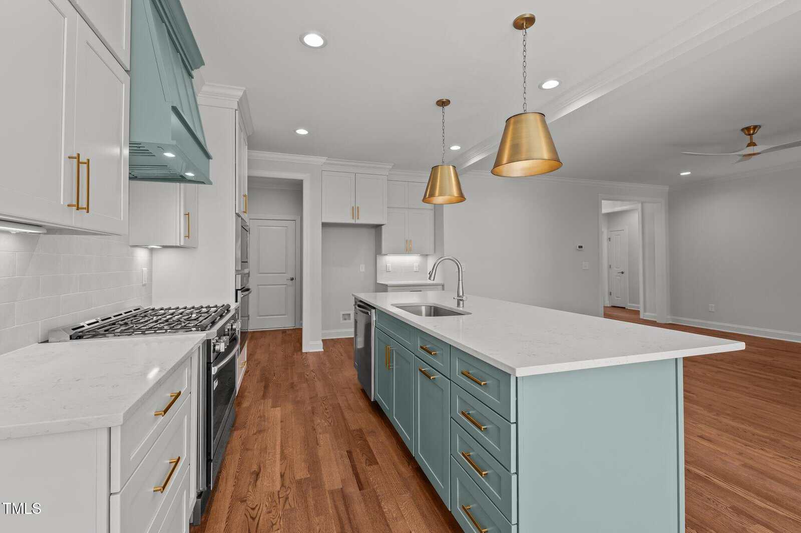 A modern kitchen with wood floors, white countertops, light blue cabinets on the island, white cabinets along the wall, stainless steel appliances, two gold pendant lights, and recessed lighting.