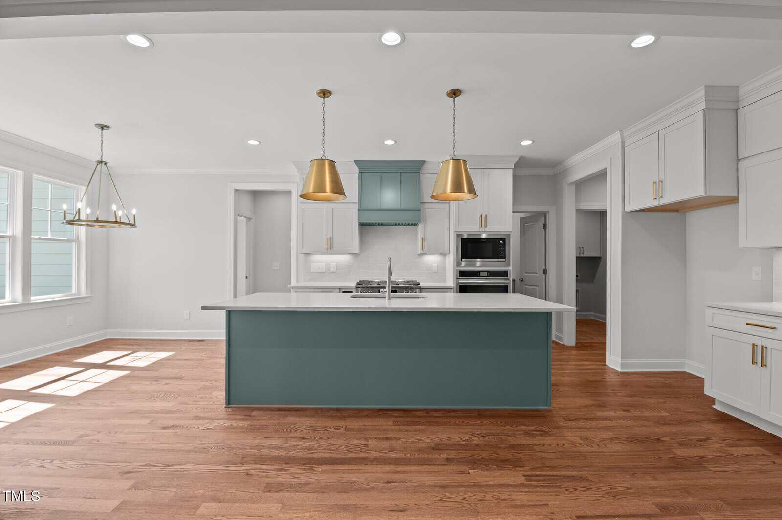 A modern kitchen with wood floors, white cabinets, a green island, gold pendant lights, a white countertop, stainless steel appliances, and a large window bringing in natural light.