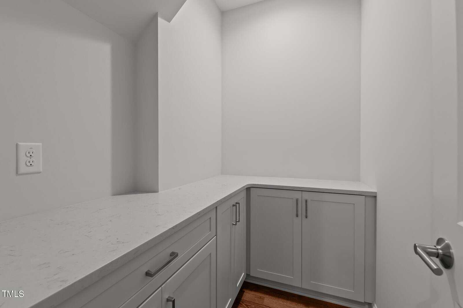 A small, modern room with white walls, gray cabinets, and a white marble countertop. There is a visible electrical outlet on the left wall and a wooden floor. The space is clean and empty.