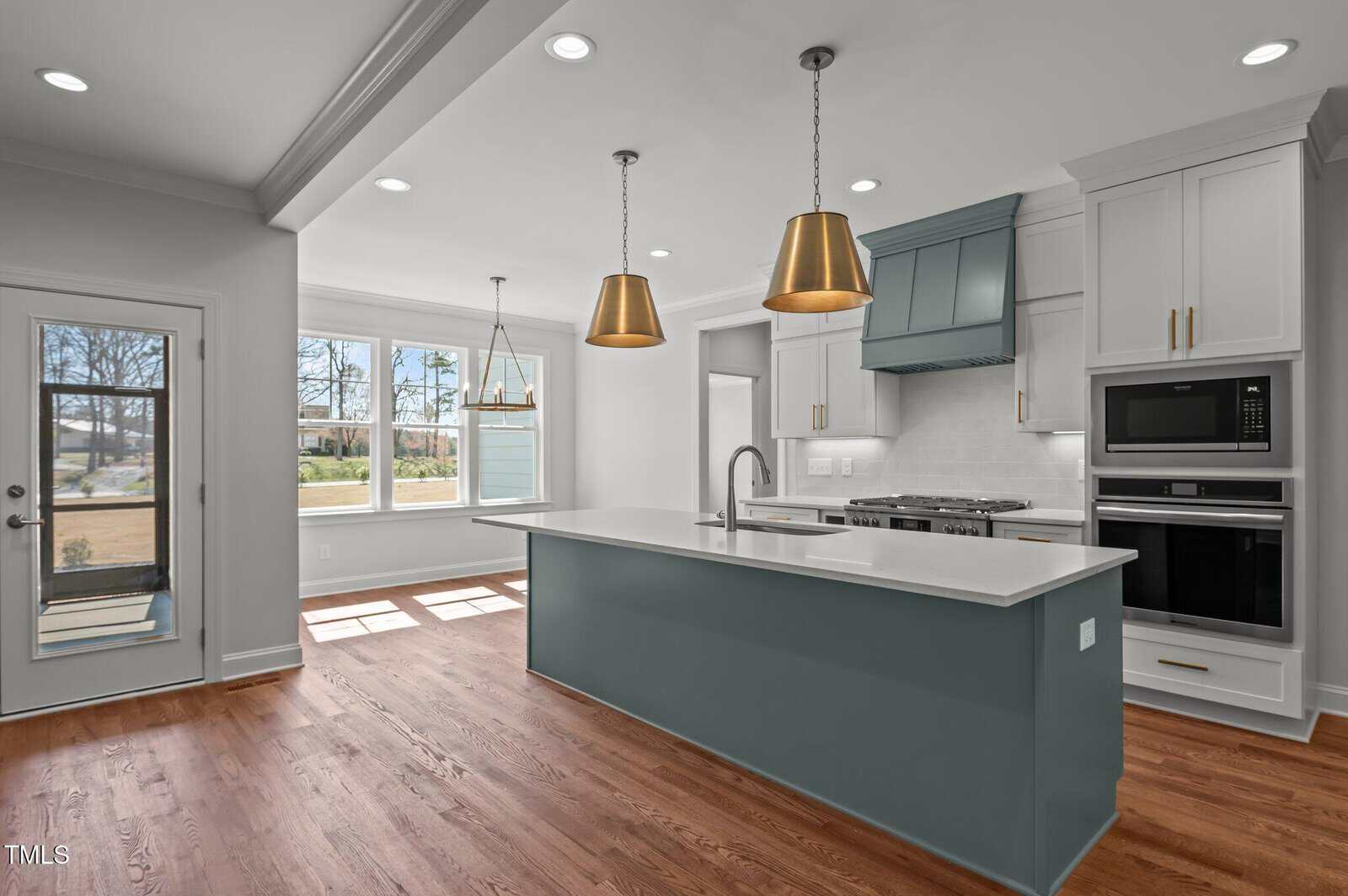 A modern kitchen with hardwood floors, a large blue-gray island with white countertops, two gold pendant lights, white cabinets, built-in oven and microwave, and a windowed breakfast nook.