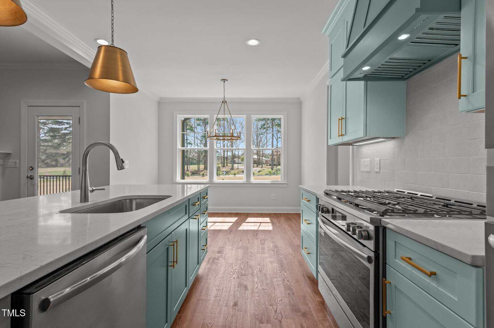 A modern kitchen features teal cabinets, stainless steel appliances, gold hardware, pendant lights, and a large window overlooking a yard. White countertops and backsplash create a clean, bright atmosphere.