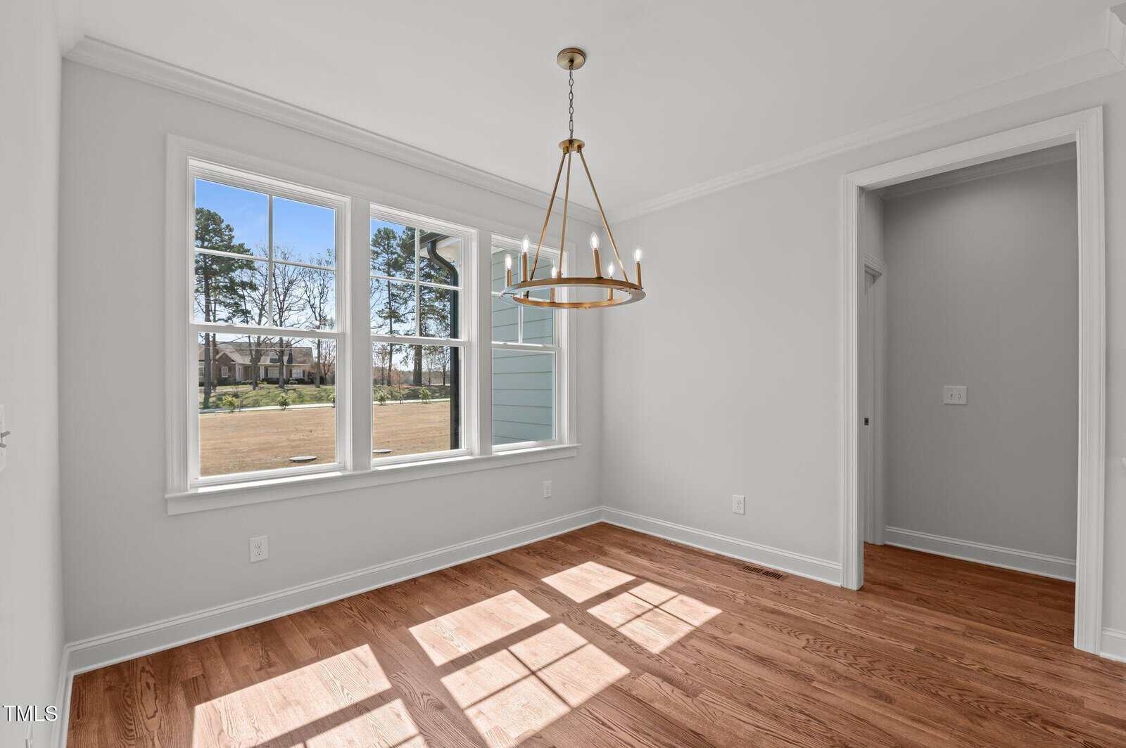 A bright, empty room with light-colored walls, a wooden floor, a large window letting in sunlight, and a modern circular chandelier hanging from the ceiling. An open doorway leads to a hallway.