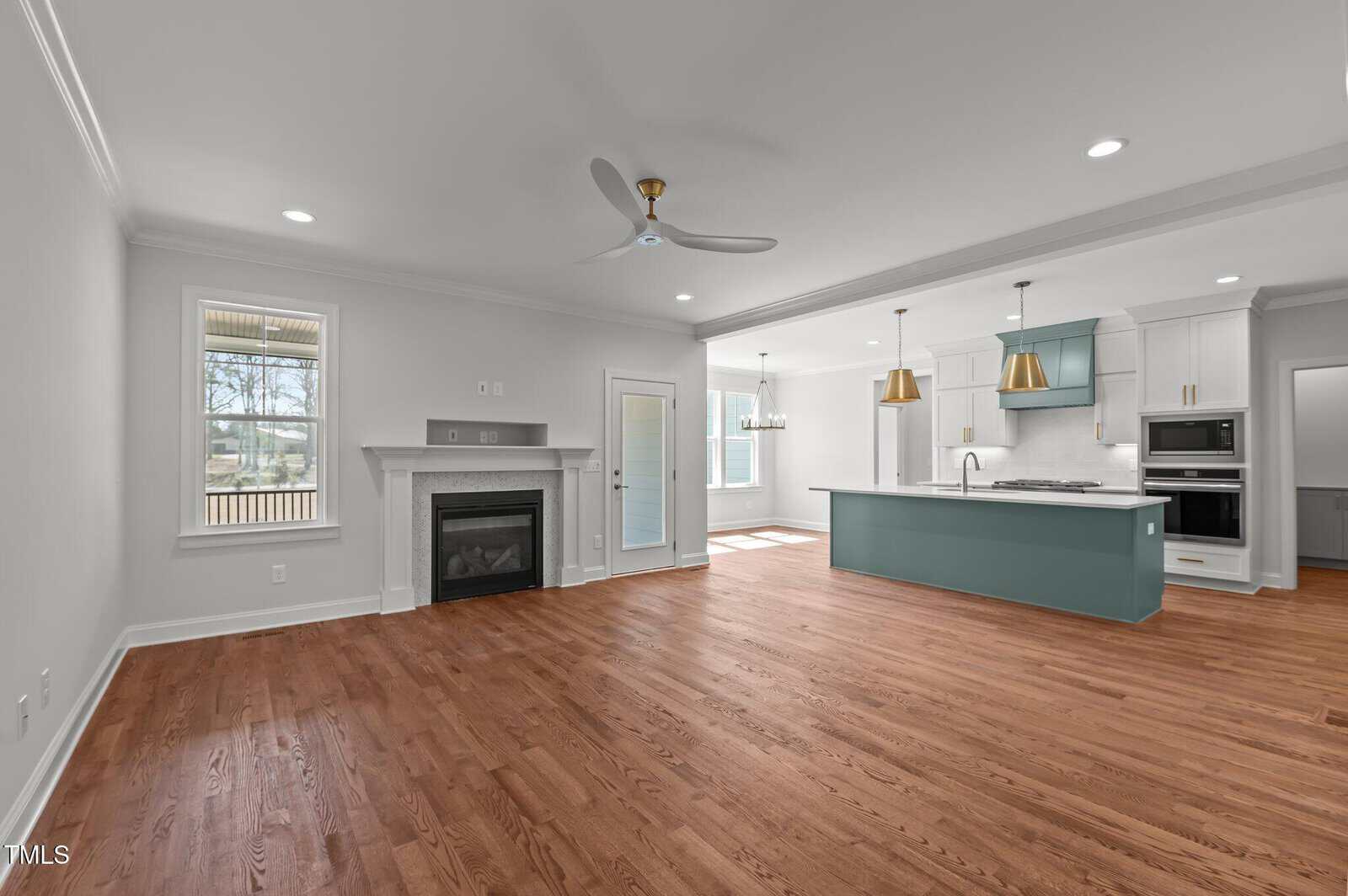 Modern open-concept living area with hardwood floors, a white fireplace, ceiling fan, large windows, and an adjacent kitchen featuring a teal island, gold pendant lights, and white cabinetry.