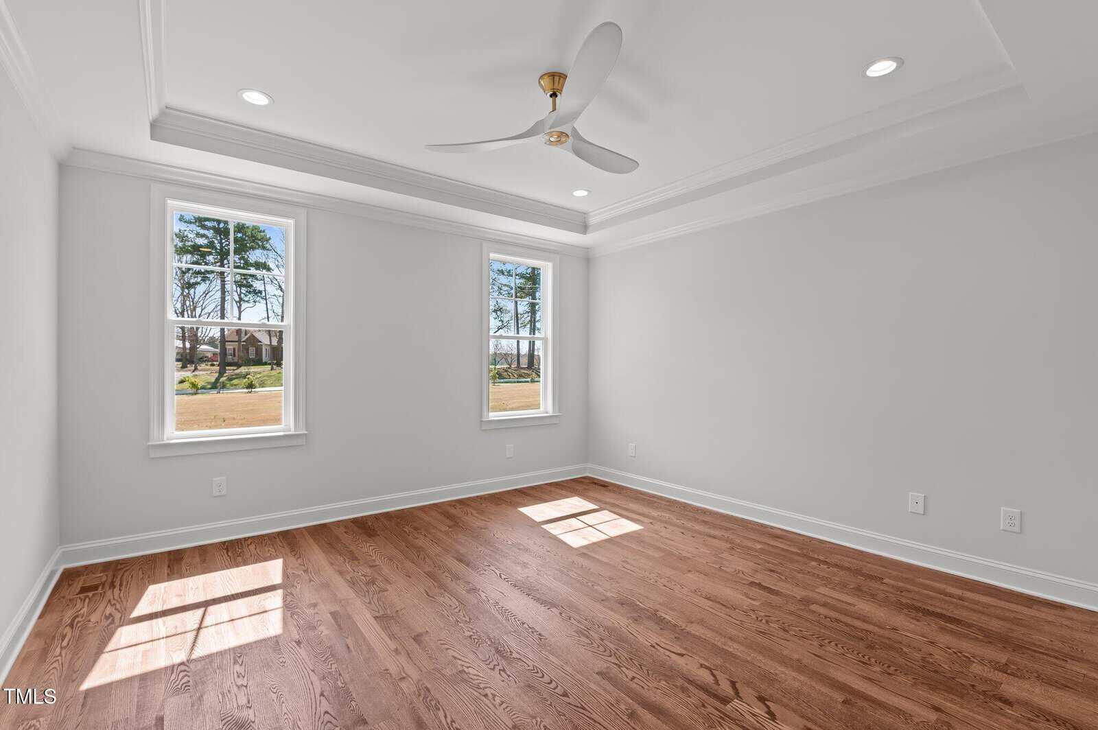 A bright, empty room with hardwood floors, two tall windows letting in natural light, white walls, recessed lighting, and a ceiling fan with a brass center.