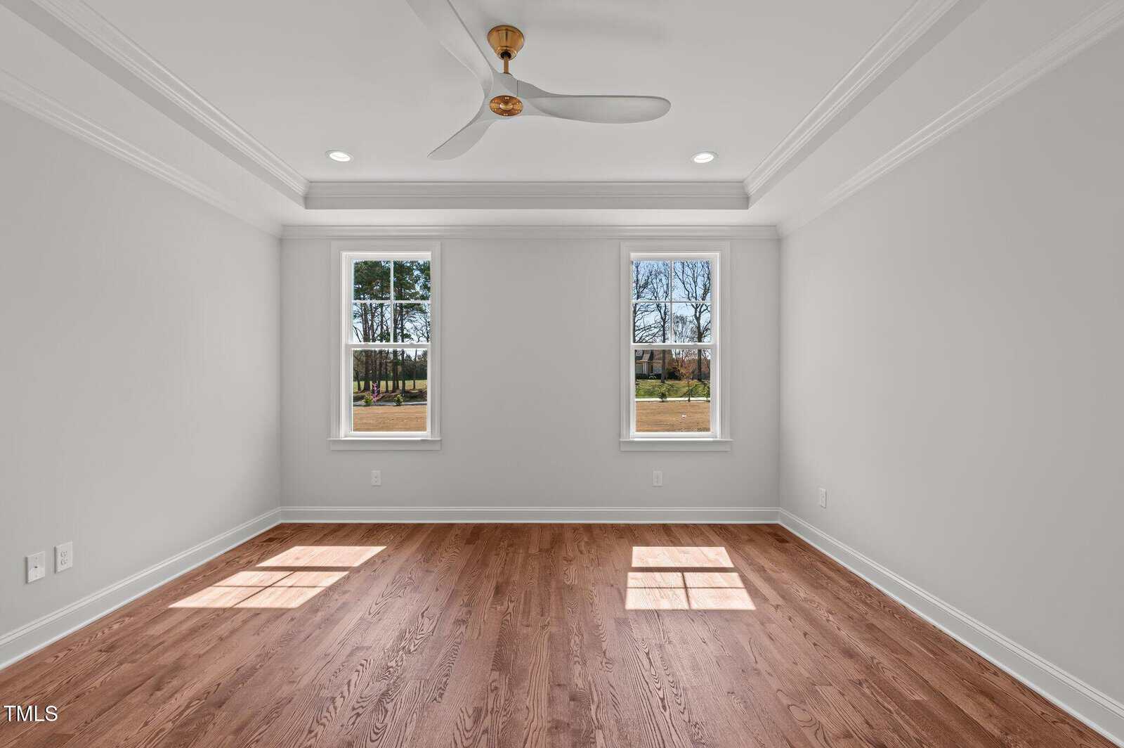 Bright, empty room with light gray walls, two large windows showing trees outside, hardwood floor, white ceiling with crown molding, recessed lights, and a ceiling fan with wooden blades.