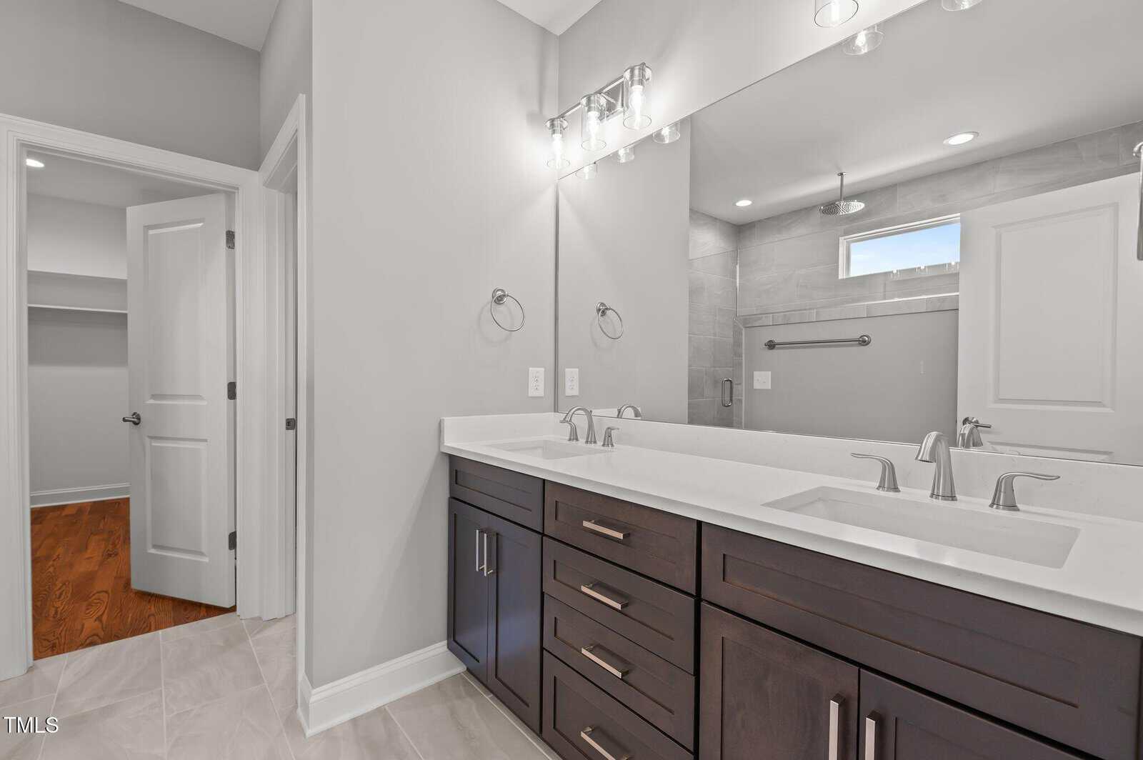 Modern bathroom with double vanity, white countertops, dark wood cabinets, large mirror, chrome fixtures, and neutral-toned tile flooring. An open door reveals a walk-in closet with wooden floors, and a glimpse of the shower.