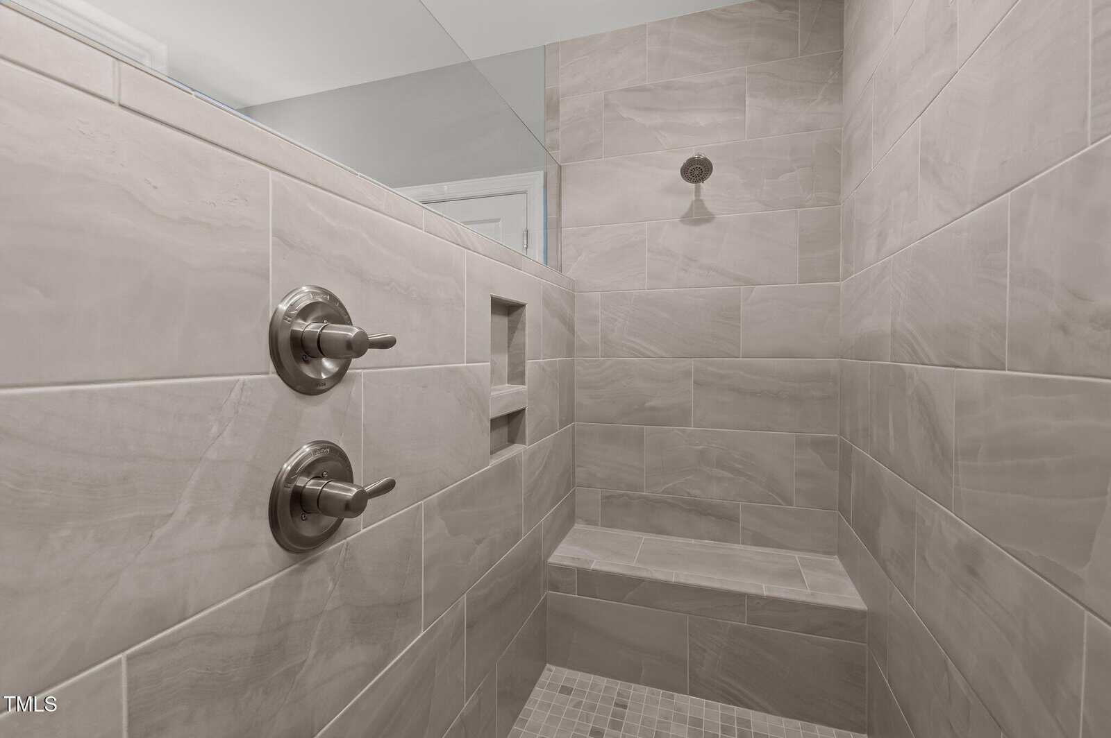 Modern walk-in shower with beige tiles, two metal control knobs, a recessed shelf, a bench, and a rainfall showerhead on the wall.