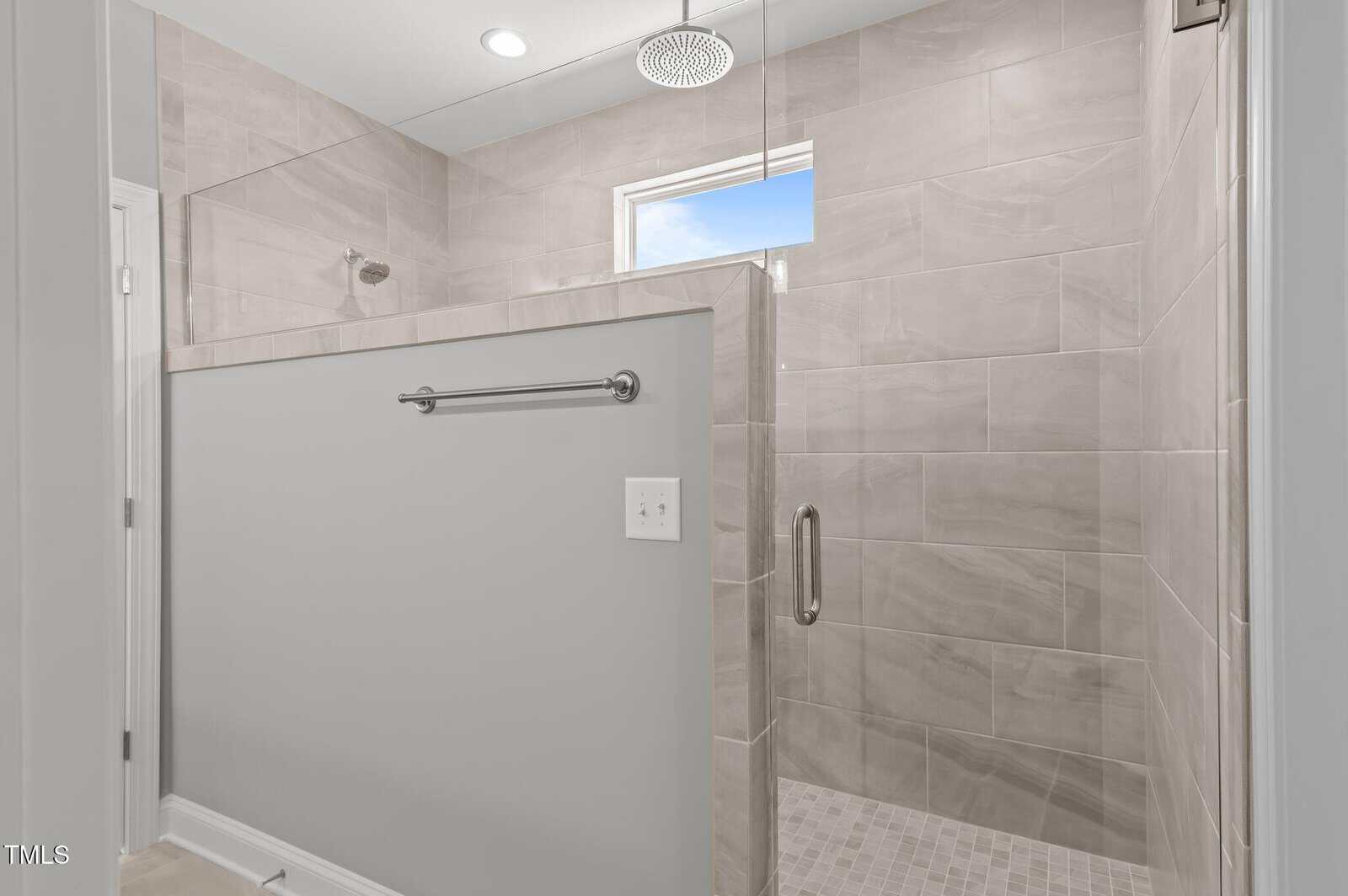 Modern walk-in shower with beige tiles, glass door, gray half wall with towel bar, rainfall showerhead, and a small window letting in natural light.