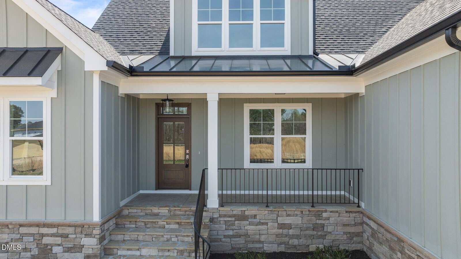 A front porch of a house with gray vertical siding, stone accents, a brown door with a glass panel, black railing, steps leading to the entrance, and large windows above and beside the door.