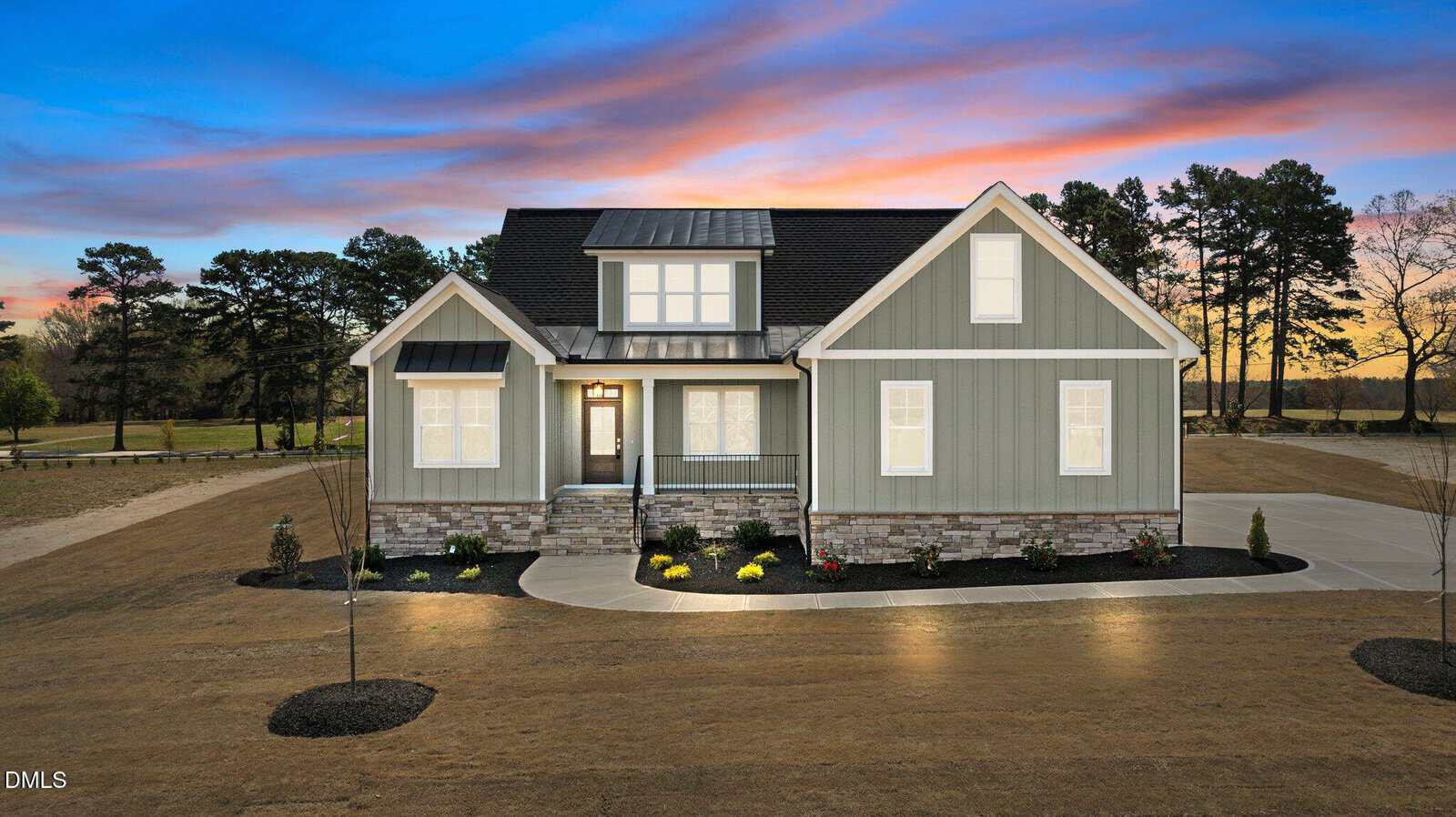 A modern gray house with white trim and stone accents is pictured at dusk, with lights on inside. The home has a covered porch, landscaped front yard, and is surrounded by trees under a colorful sunset sky.