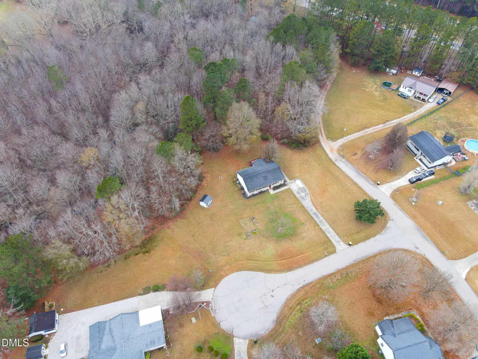 Aerial view of a suburban cul-de-sac bordered by a wooded area. Several houses with lawns and driveways are visible, one with a shed and garden beds. The landscape is mostly brown with scattered trees.