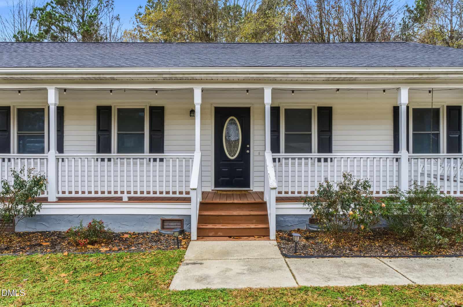 A white single-story house with a dark shingled roof, central black front door with oval window, wooden steps, and a wide front porch with railings. Small bushes line the front and trees are visible in the background.