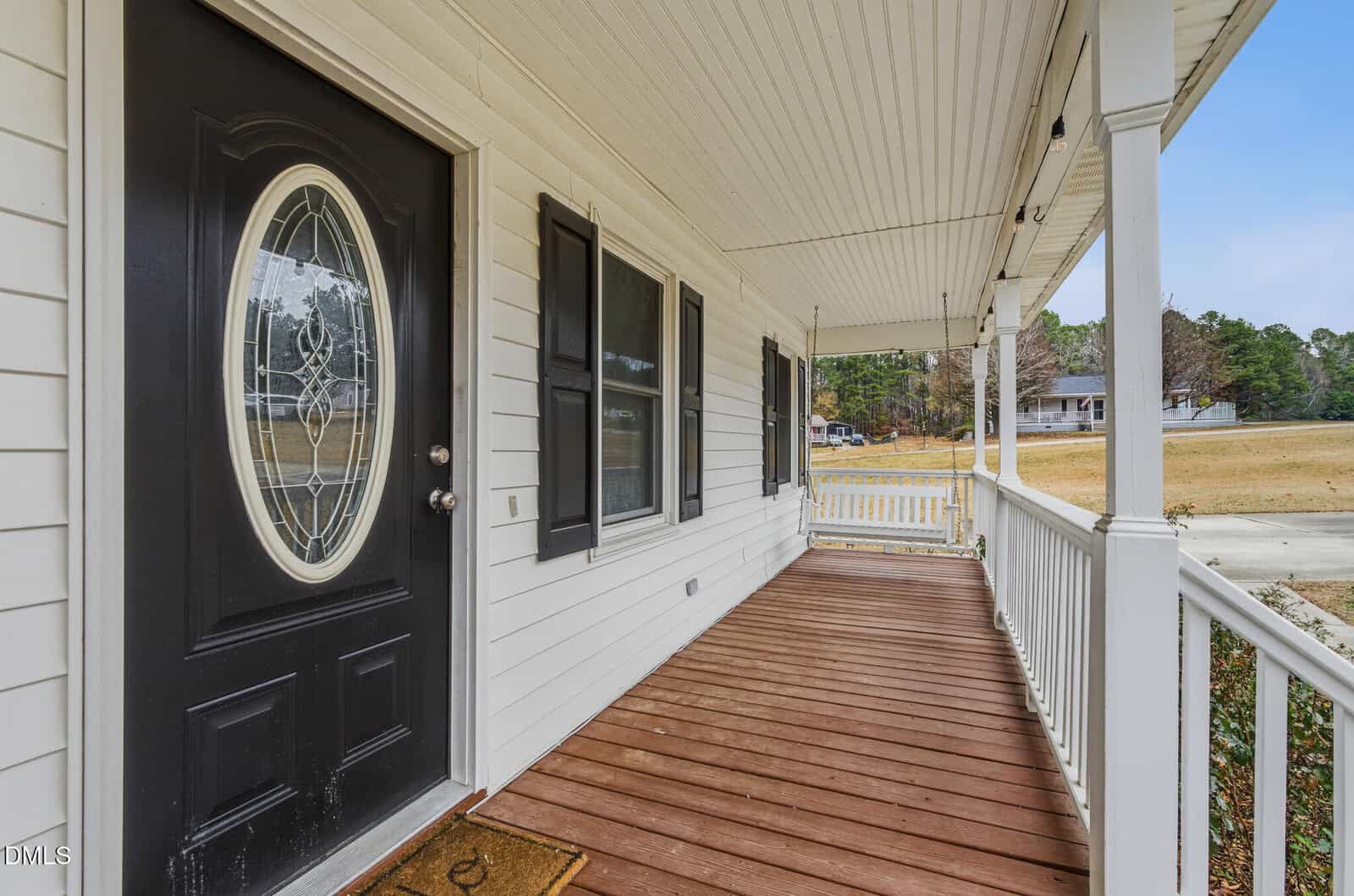 A front porch with white railings and siding, brown wooden floorboards, two black shuttered windows, and a black door with an oval glass insert, overlooking a grassy yard and distant houses.