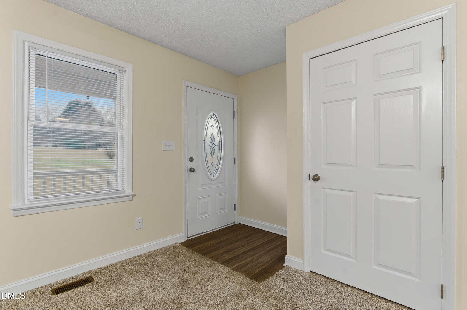 A small entryway with a white front door featuring an oval glass window, a white closet door, beige walls, a window with blinds, and a mix of carpet and wood flooring.