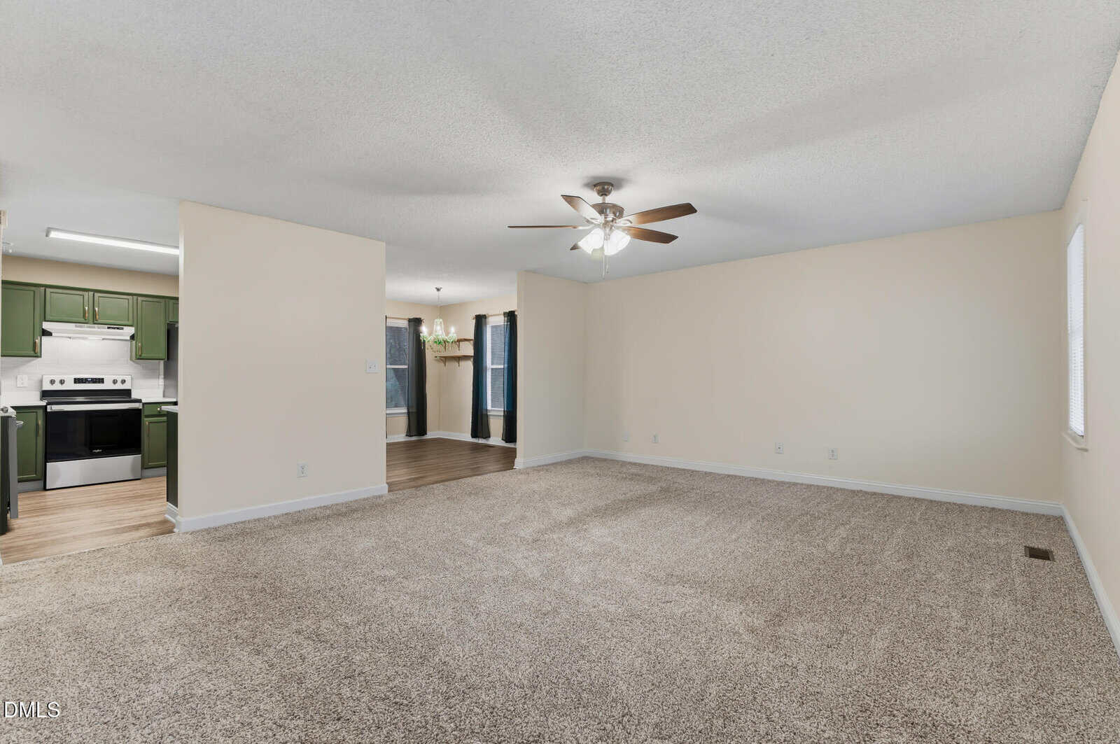Spacious living room with beige carpet, light-colored walls, and a ceiling fan with lights. The room opens to a kitchen with green cabinets and stainless steel appliances, and a dining area with wood flooring.